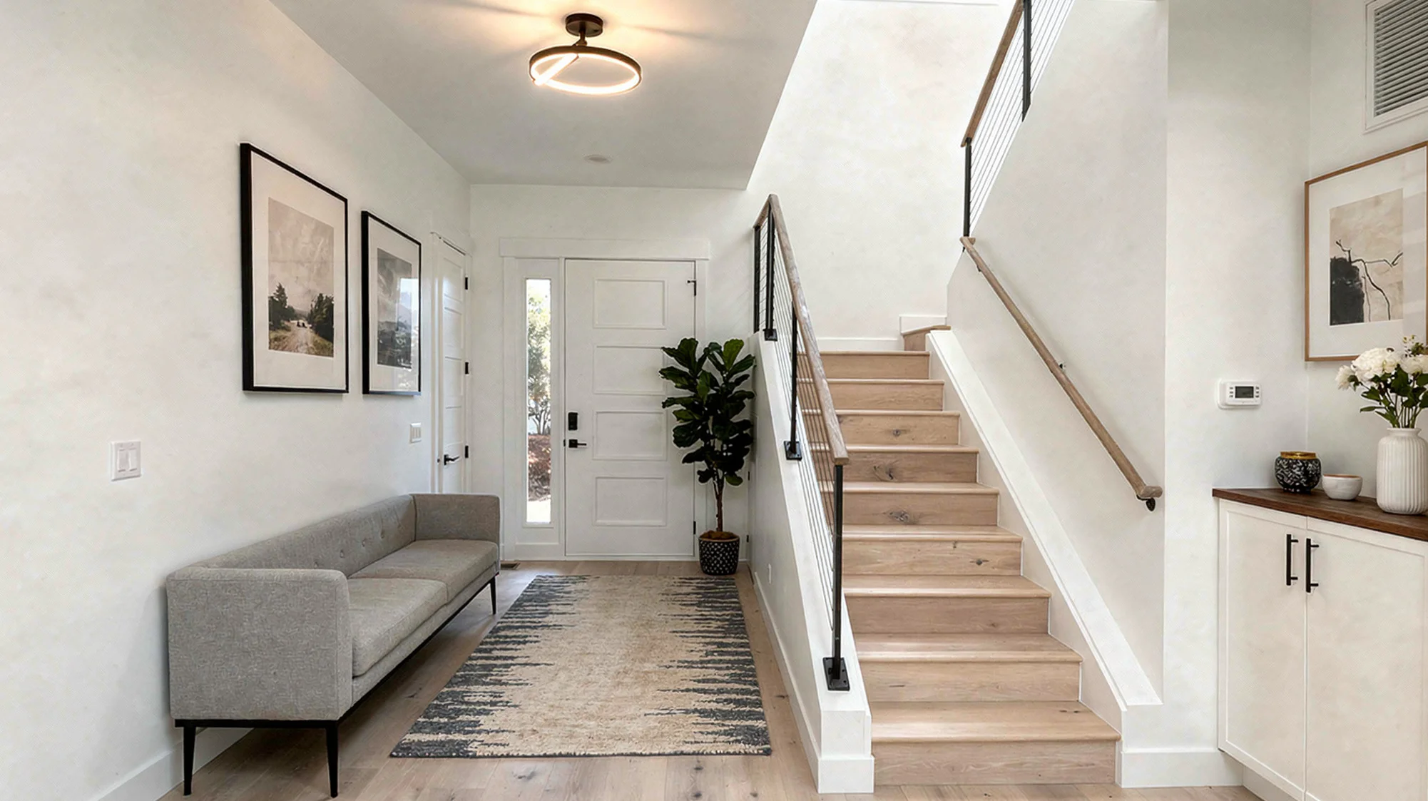 Sunlight entry foyer with gray settee, light wood staircase with cable railing, fiddle-leaf fig, and framed art on white walls