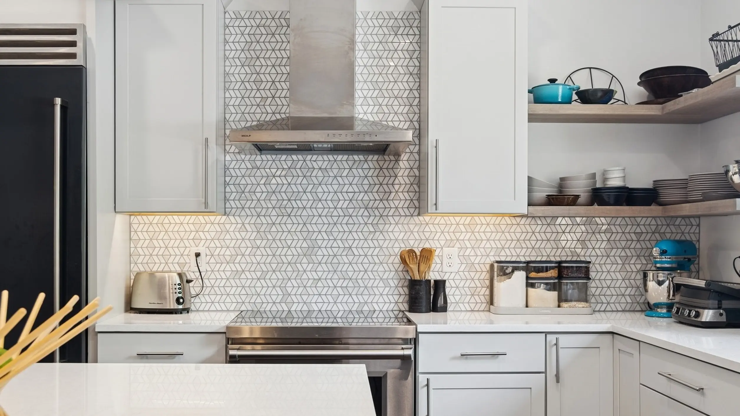 Skyview kitchen backsplash detail with geometric patterned tile, white shaker cabinets, open shelving with cookware, and stainless range