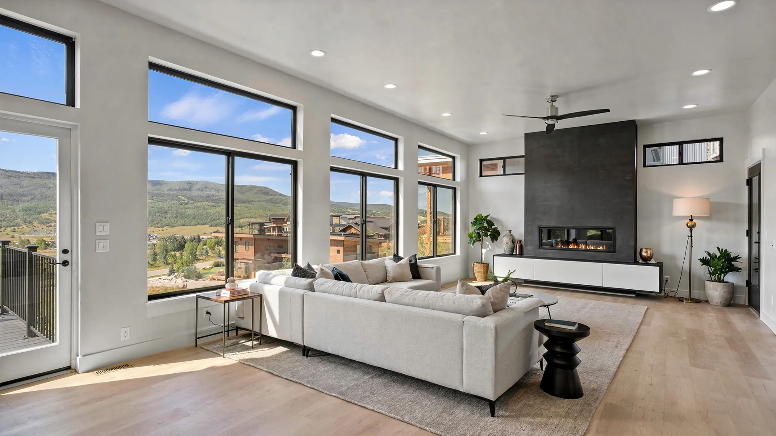 Sunlight living room with cream sectional, dark slate fireplace feature wall with linear fireplace, ceiling fan, and large windows with mountain and neighborhood views