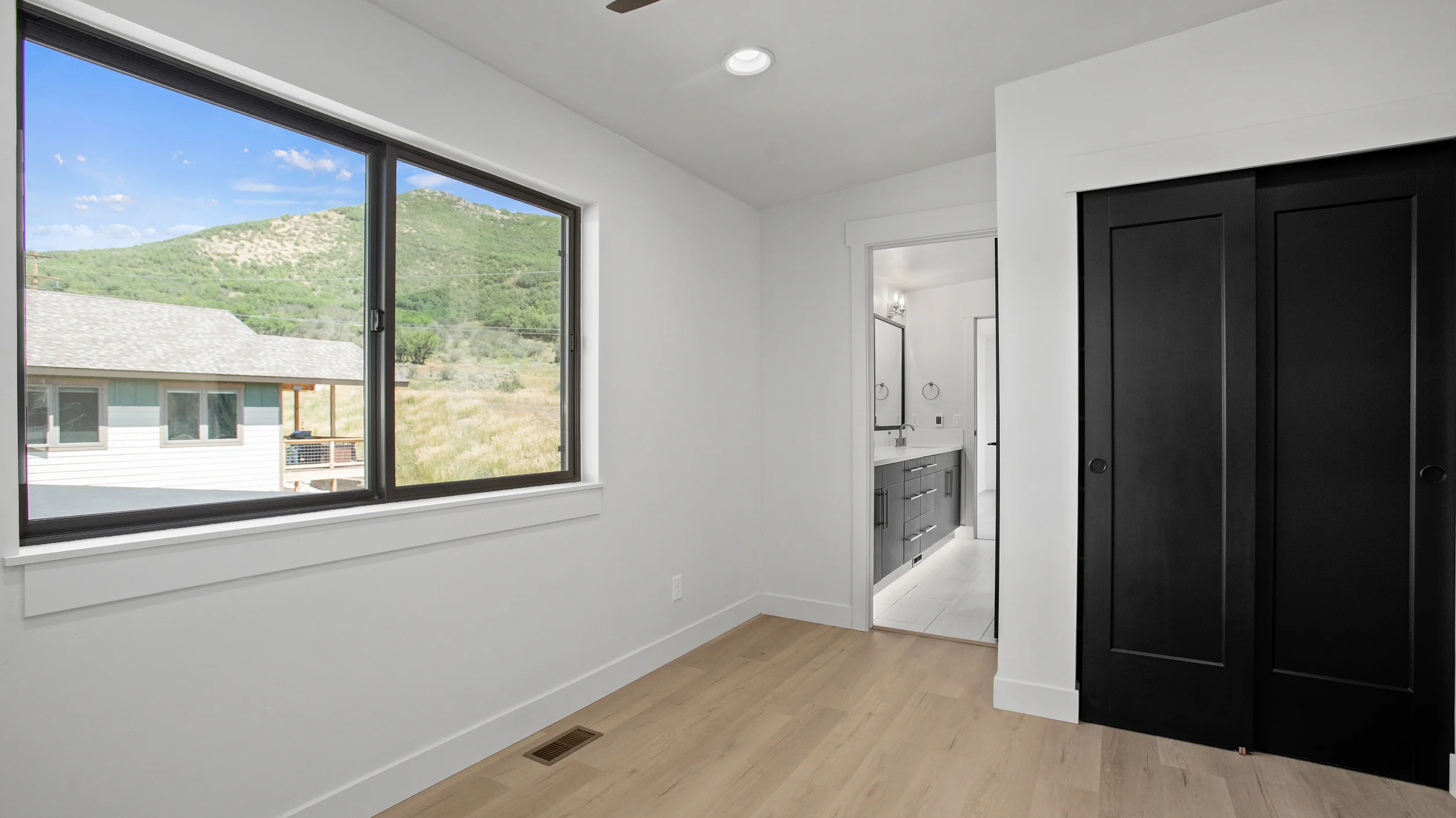 Sunlight bedroom with large window showing green mountain views, black double closet doors, and en suite bathroom doorway