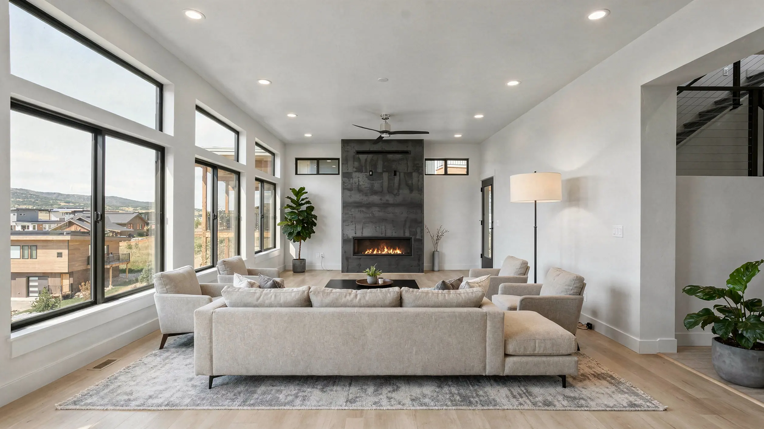 Sunlight upper-level great room with large gray L-shaped sectional, dark slate fireplace accent wall, linear fireplace, ceiling fan, and floor-to-ceiling windows with mountain and neighborhood views
