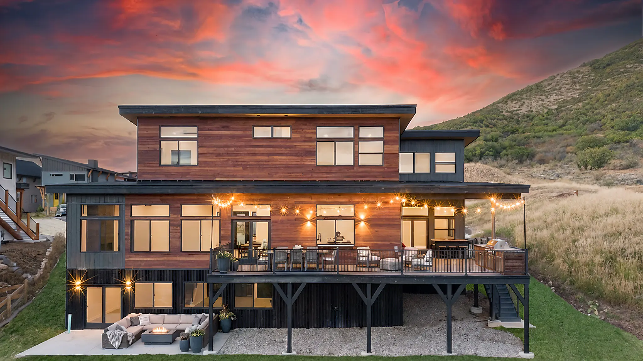 Sunlight rear exterior at vivid sunset showing two-story home with cedar siding, wide upper deck with string lights and outdoor dining, lower patio with sofa, and green hillside