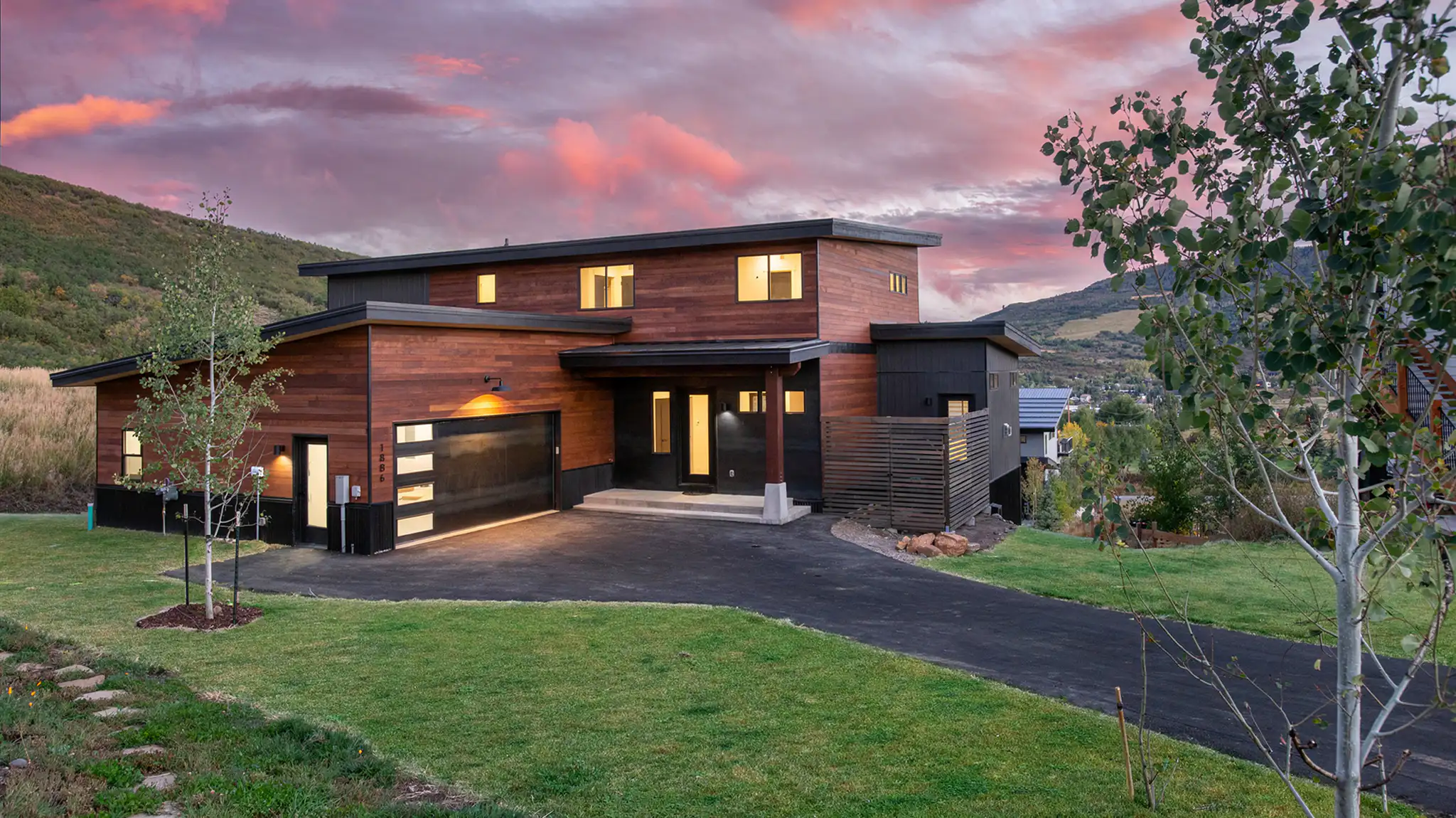 Sunlight front exterior at dusk with cedar siding, dark lower level and trim, attached 2-car garage, lit windows, and mountain hillside with aspens behind