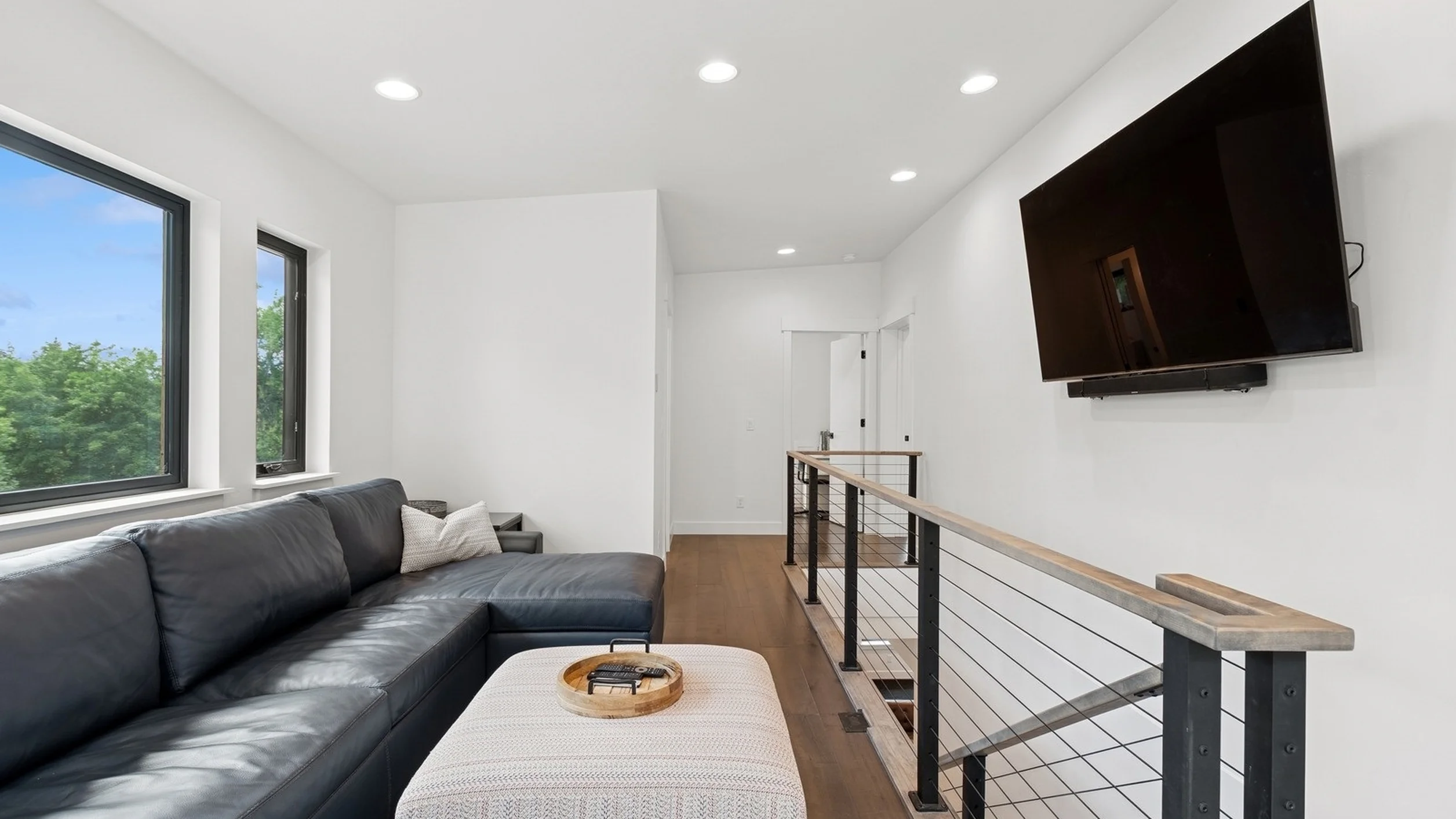 Skyview upper-level bonus room with dark leather sectional, wall-mounted TV, and open cable railing at staircase with treetop views