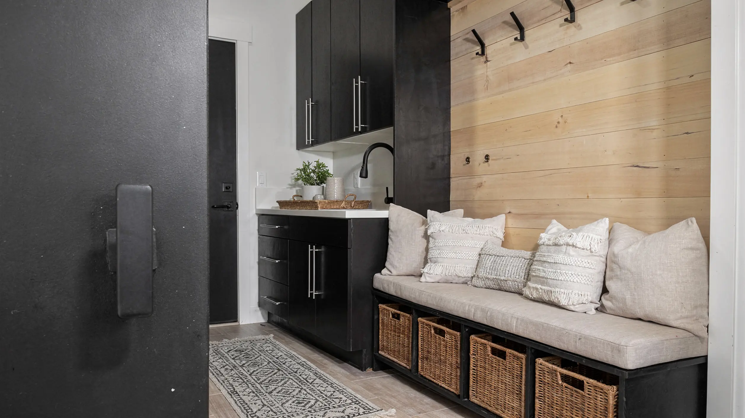 Sunlight mudroom with cedar shiplap bench with cushioned seat and wicker storage baskets, black upper cabinets with sink, and wall hooks for coats