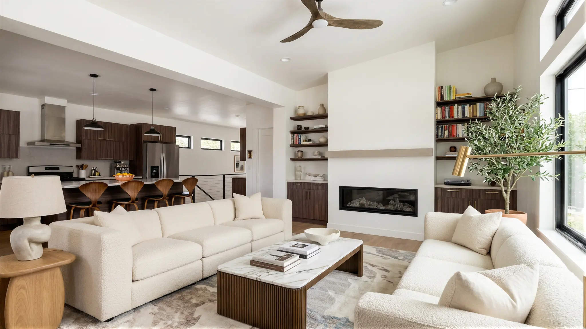 Bluebird open-plan great room with cream sofas, linear fireplace, floating shelves with books, ceiling fan, and dark wood kitchen with island visible beyond