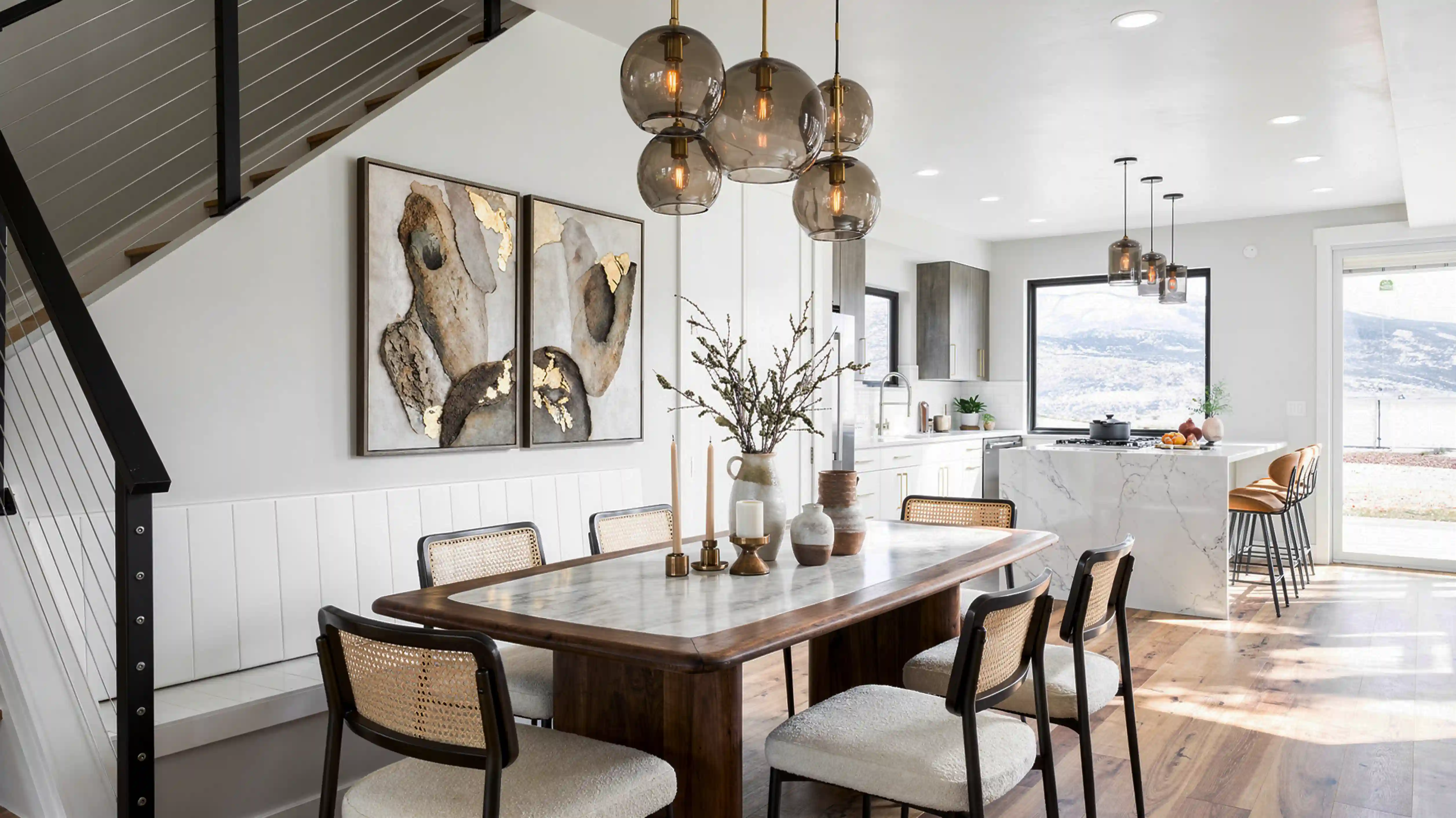 Twilight dining area with marble-top table, cane-back chairs, globe cluster pendant lights, and open kitchen with mountain views beyond