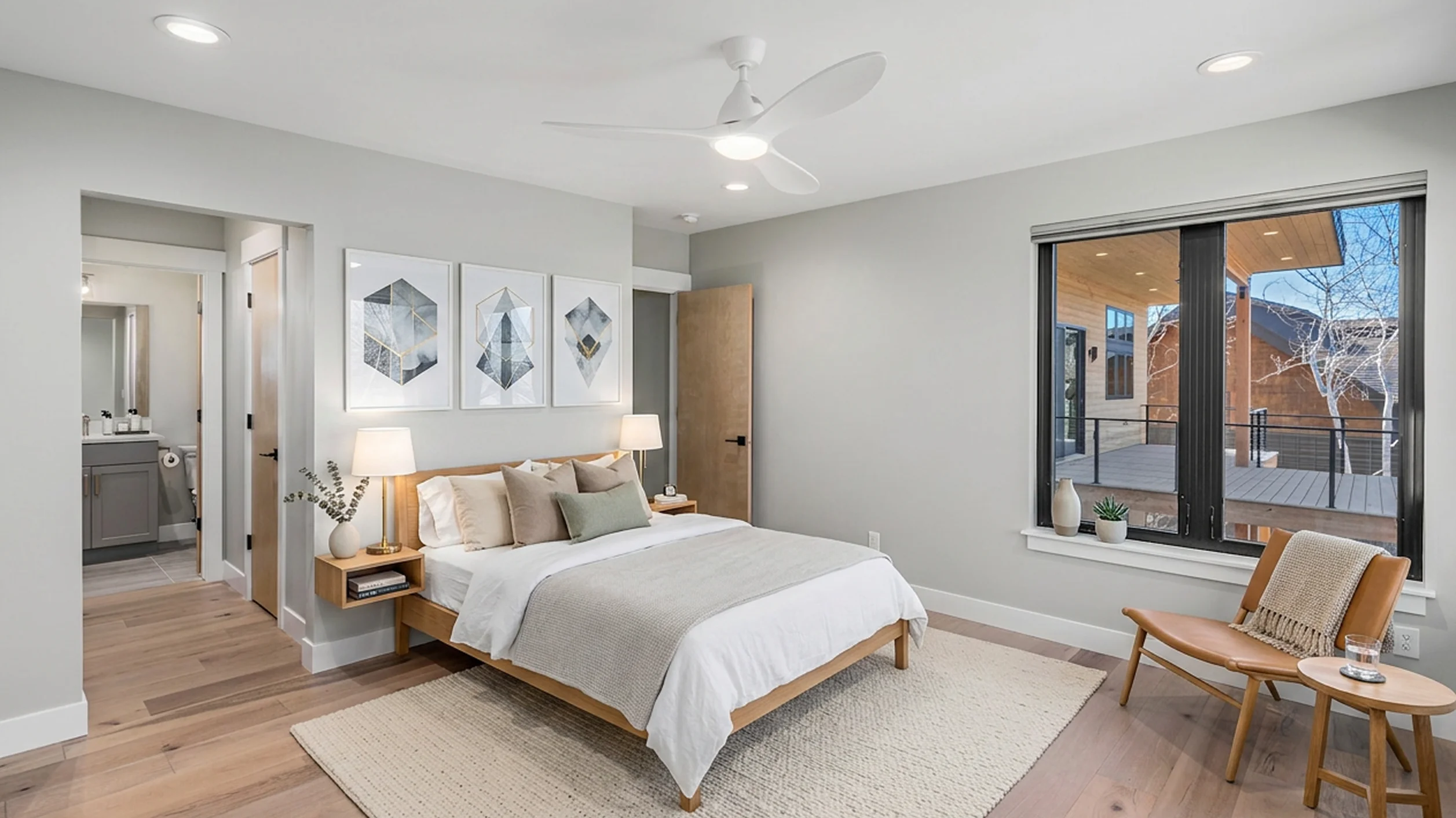Telemark primary bedroom with wood-frame platform bed, ceiling fan, geometric art triptych, window overlooking deck railing, and en suite bathroom doorway