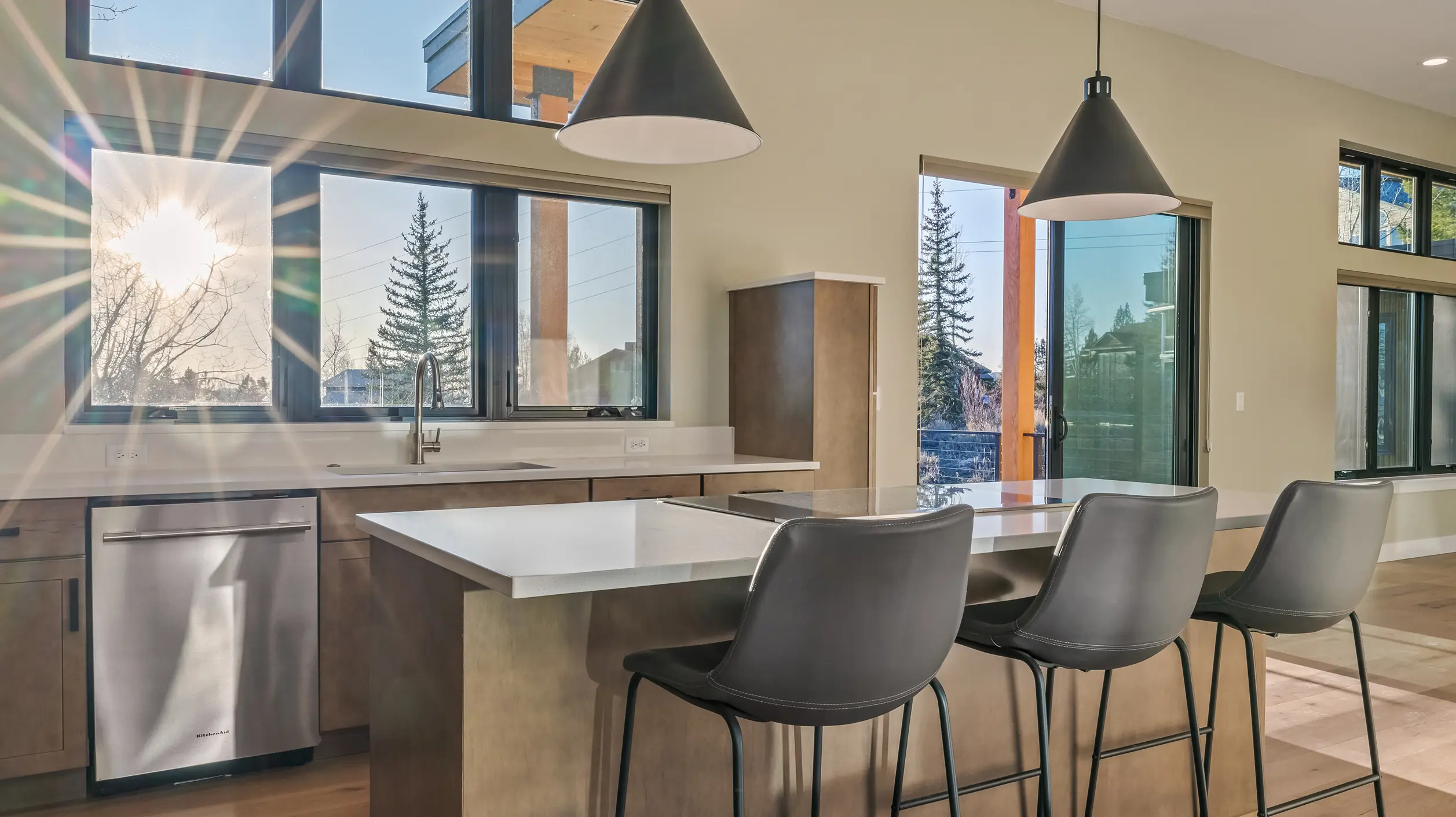 Telemark kitchen with wood cabinetry, white quartz peninsula with gray barstools, black cone pendants, and large windows with pine tree and neighborhood views