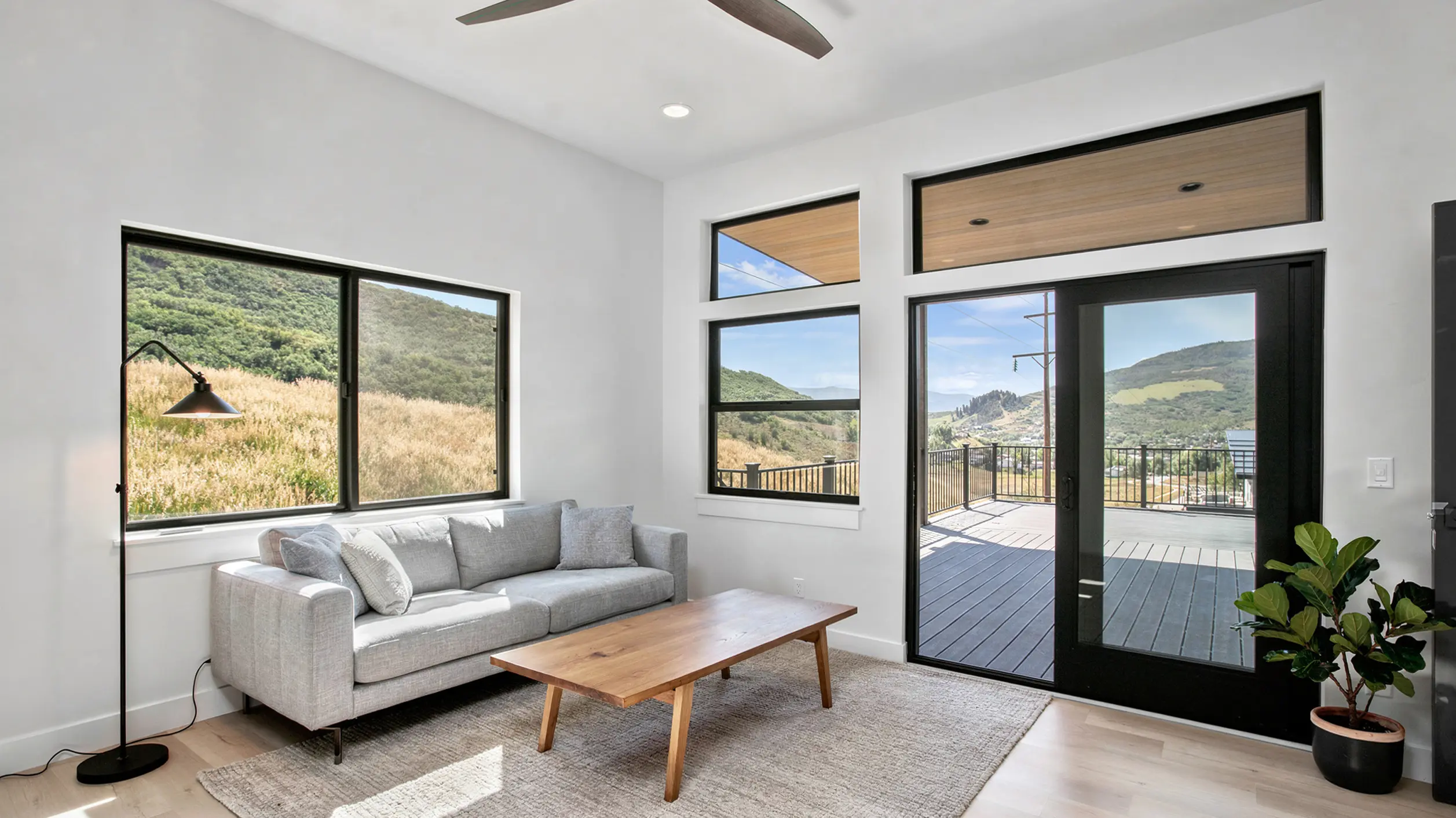 Sunlight lower-level sitting room with gray sofa, wood coffee table, ceiling fan, floor-to-ceiling windows with green mountain views, and sliding glass door to deck