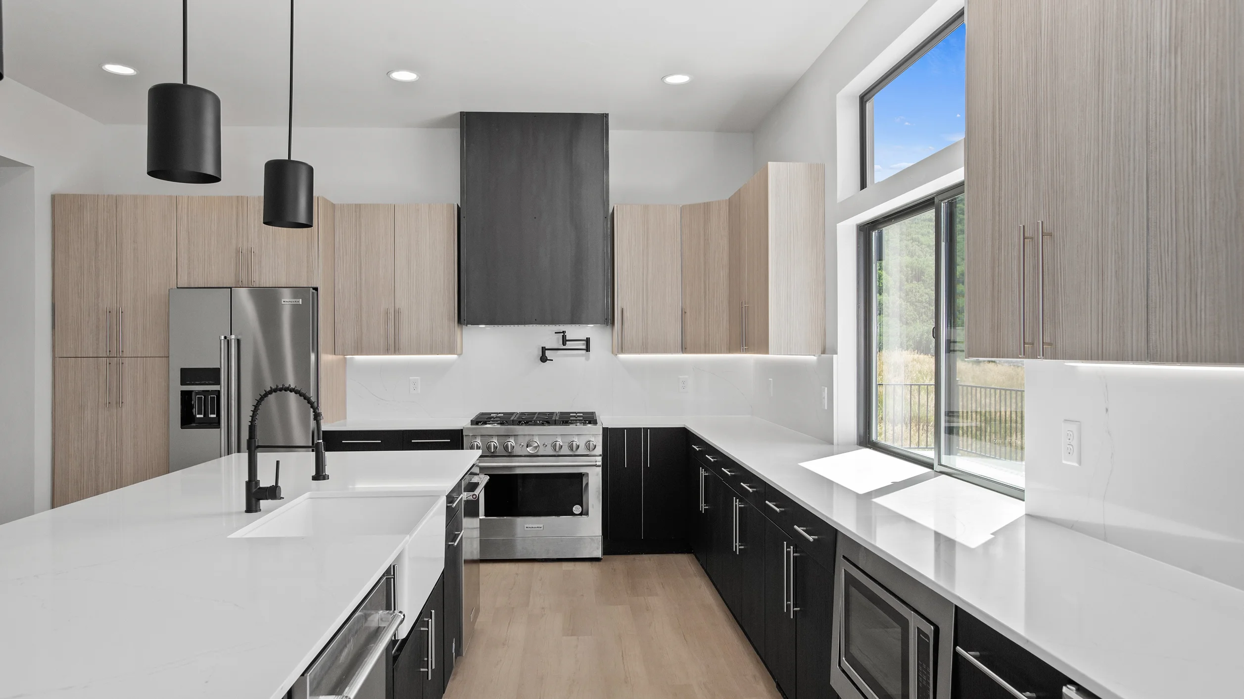 Sunlight kitchen with two-tone cabinetry, white quartz island with sink, stainless range and dark custom hood, black cylinder pendants, and window with deck view