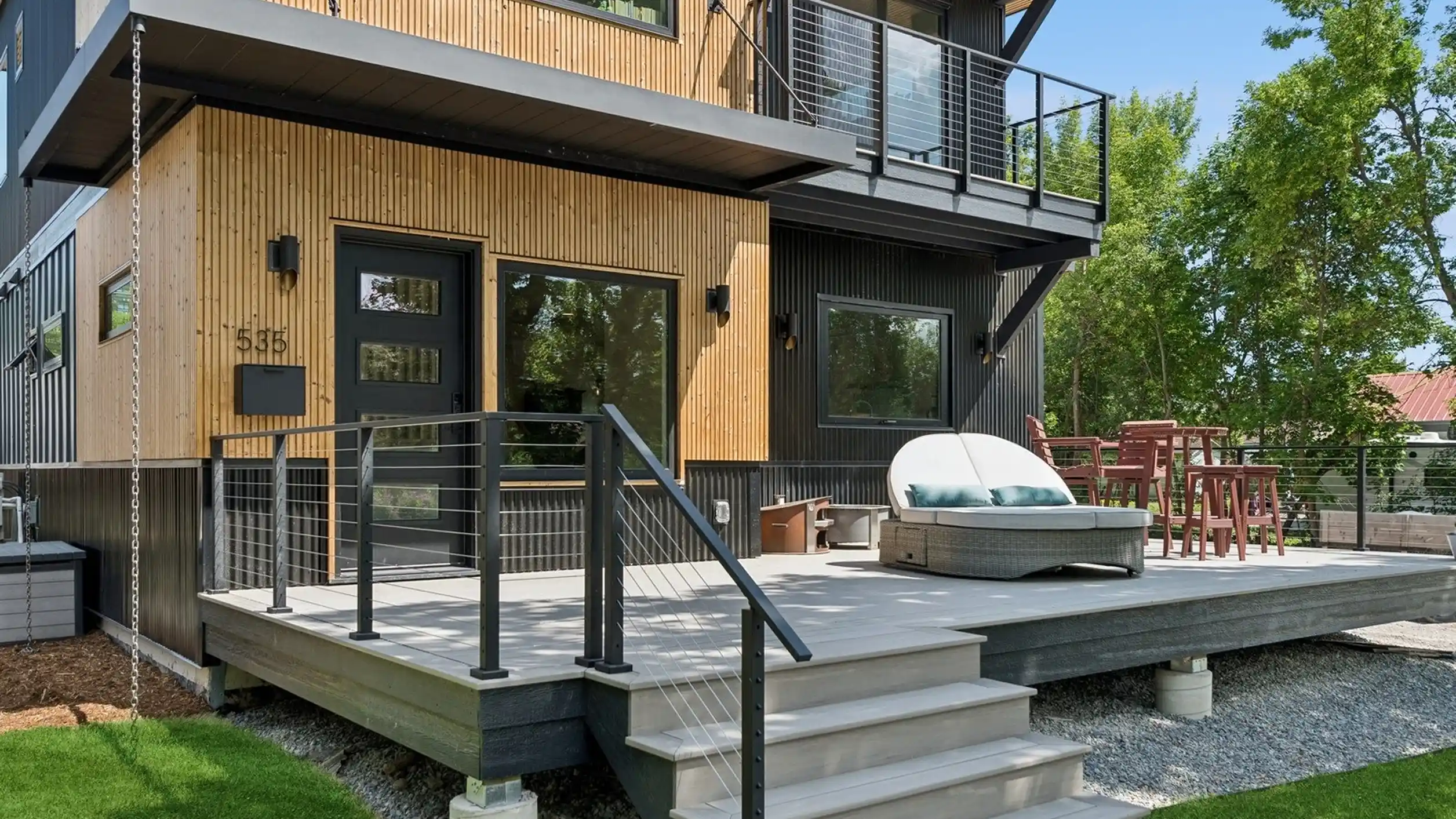Skyview exterior rear view showing dark board-and-batten and wood siding, ground-level deck with lounge chair, upper deck with cable railing, surrounded by mature trees
