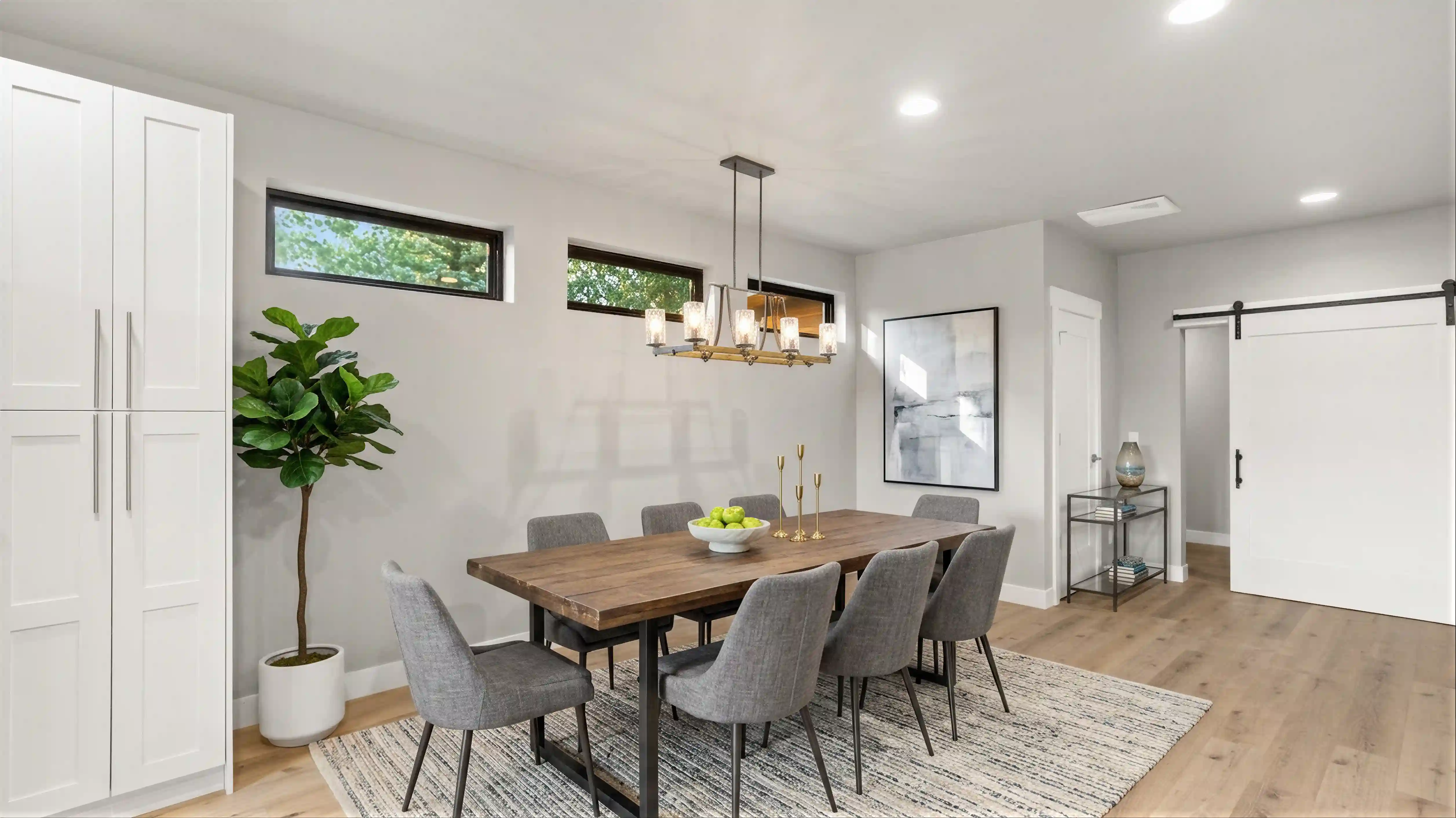 High Noon dining room with wood plank table, gray upholstered chairs, linear brass chandelier, sliding barn door, and clerestory windows