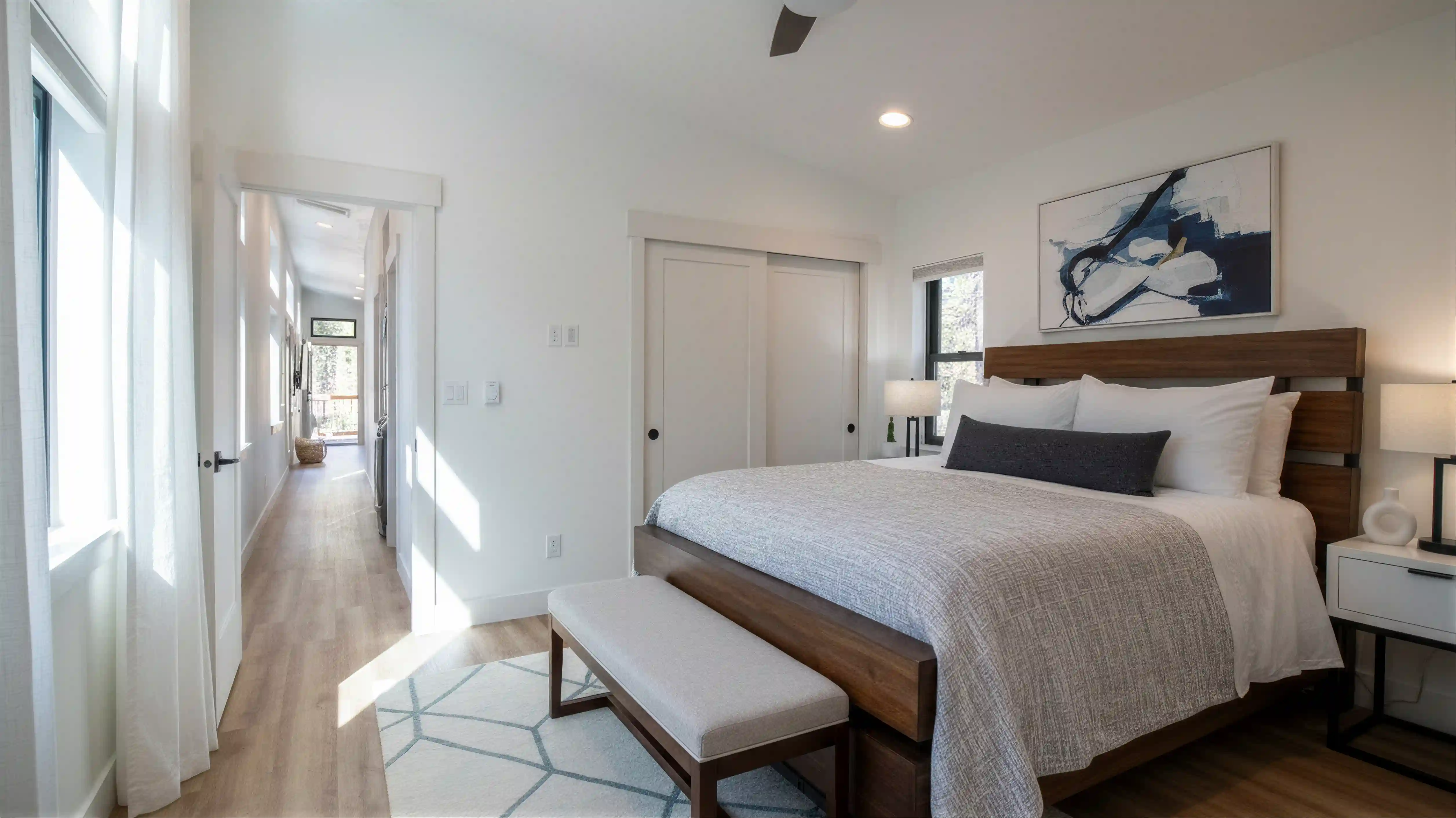Hideaway bedroom with wood-frame bed, end-of-bed bench, abstract art above headboard, ceiling fan, and sunlit hallway visible to the left