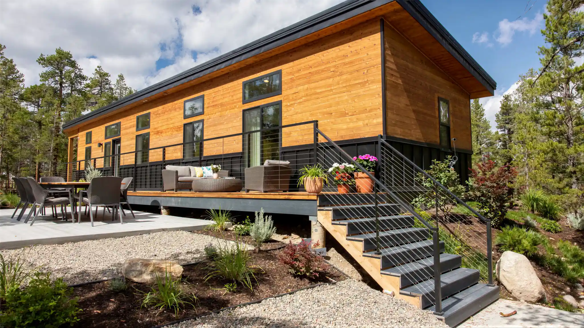 Hideaway exterior side view with cedar siding, deck featuring outdoor sofa and dining furniture, cable railing, stairs, and pine forest backdrop