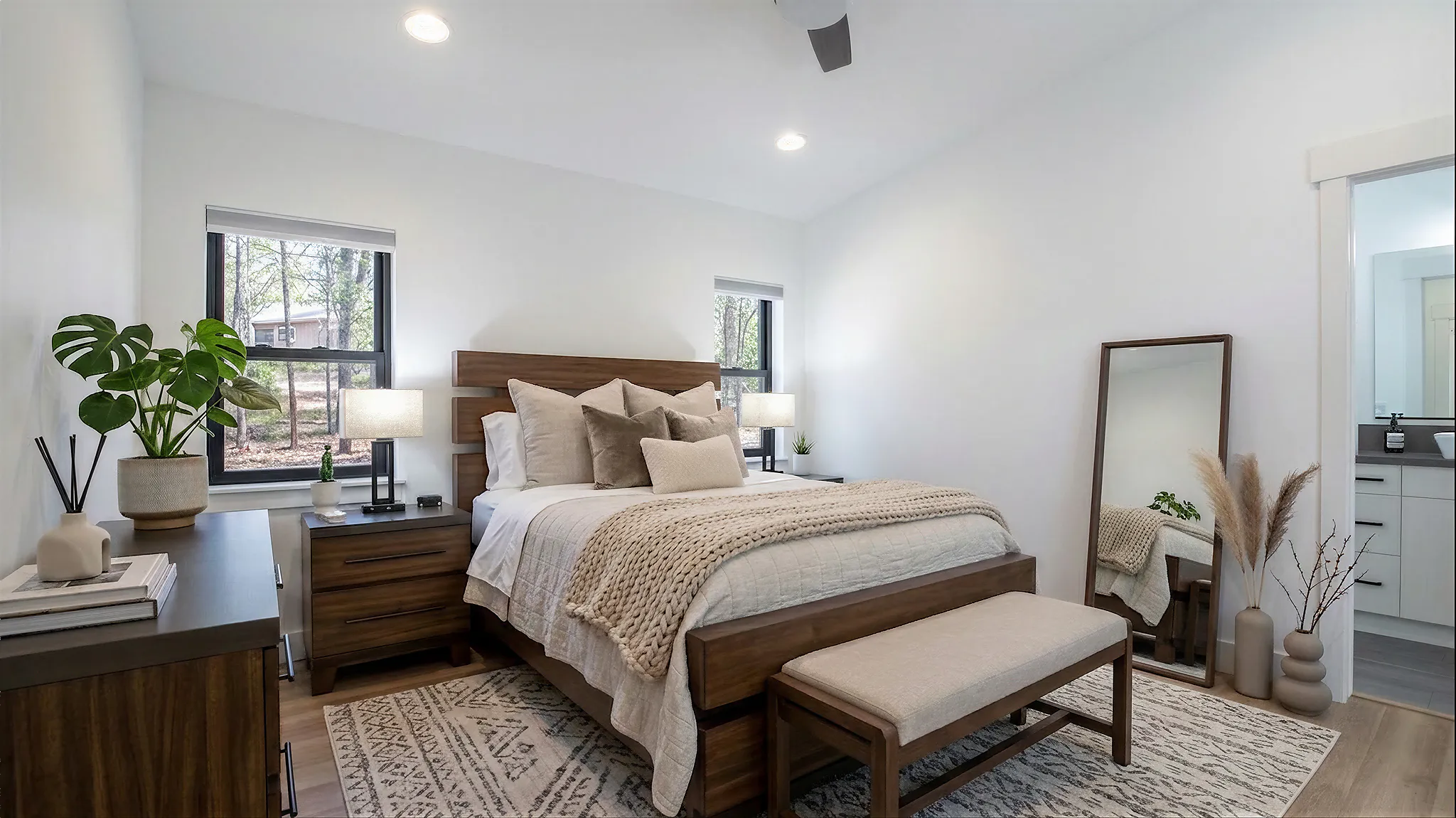 Hideaway spacious primary bedroom with wood-frame bed, matching nightstands, bench at foot, houseplants, full-length mirror, and en suite bathroom doorway