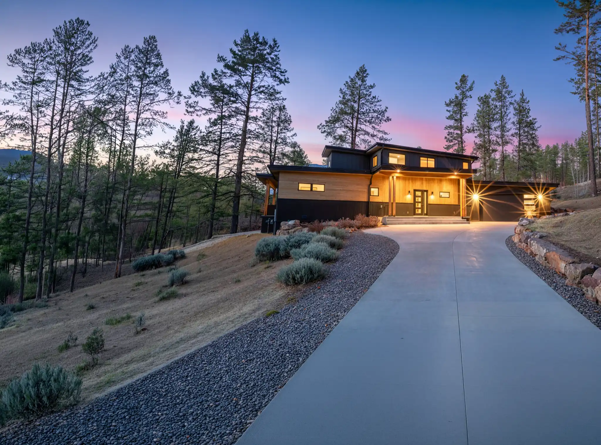 Two-story home with cedar and dark siding on a pine-covered hillside, with a long concrete driveway and vivid pink sunset sky