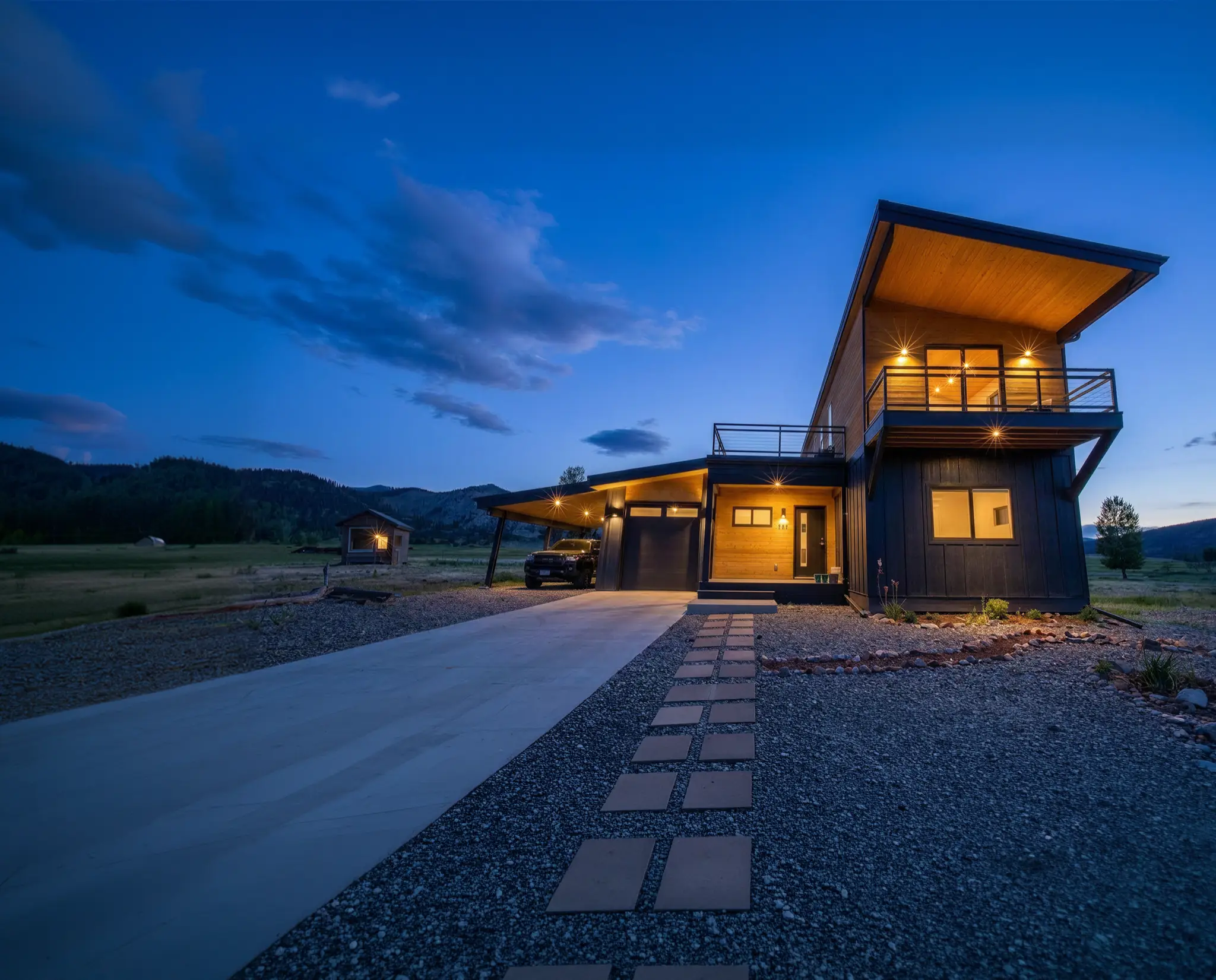 Two-story home with dark vertical siding and cedar-lined upper deck, set in an open rural valley with rolling hills at dusk