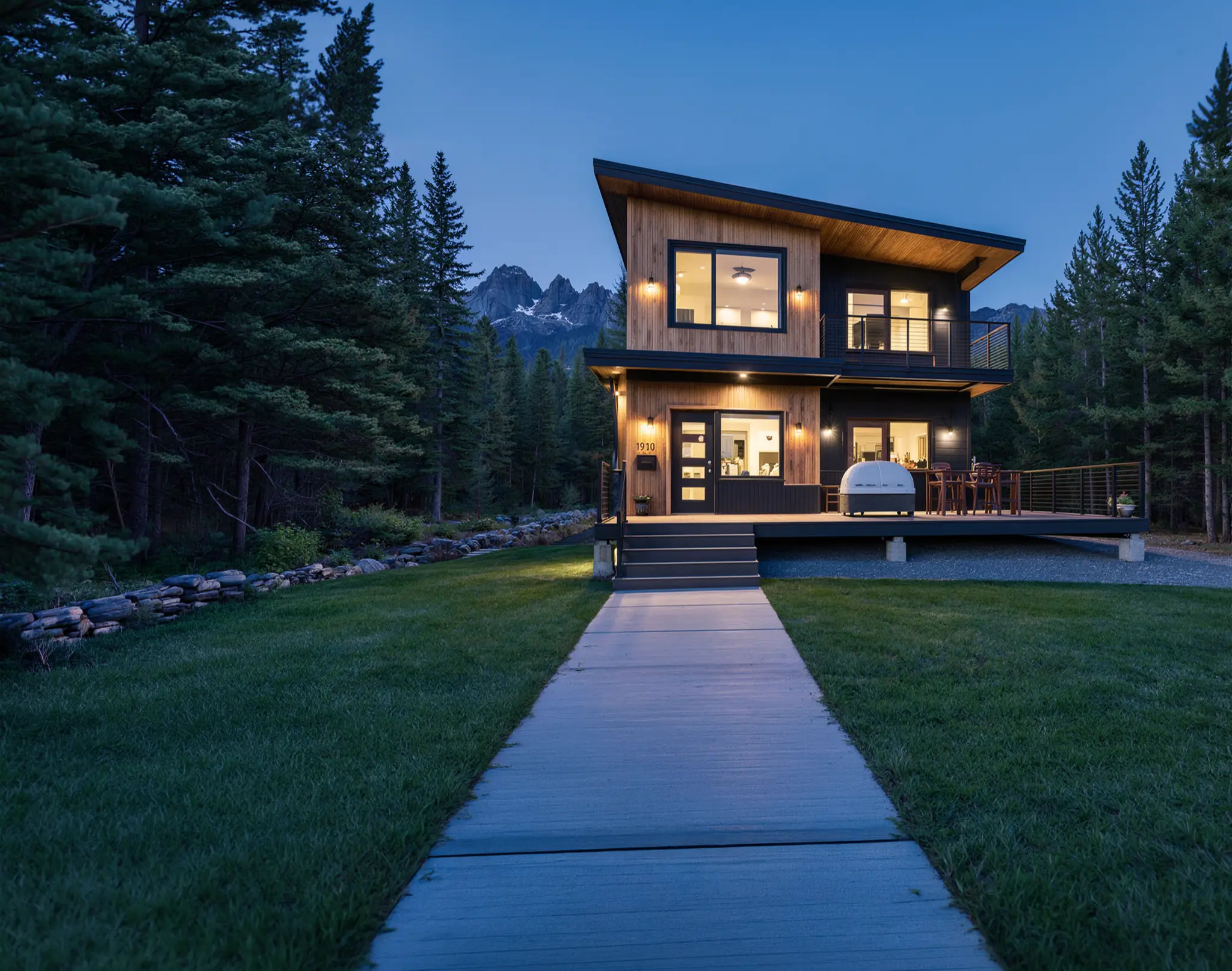 Two-story mountain home with cedar and dark siding, connected by a boardwalk path through a green lawn with jagged peaks behind