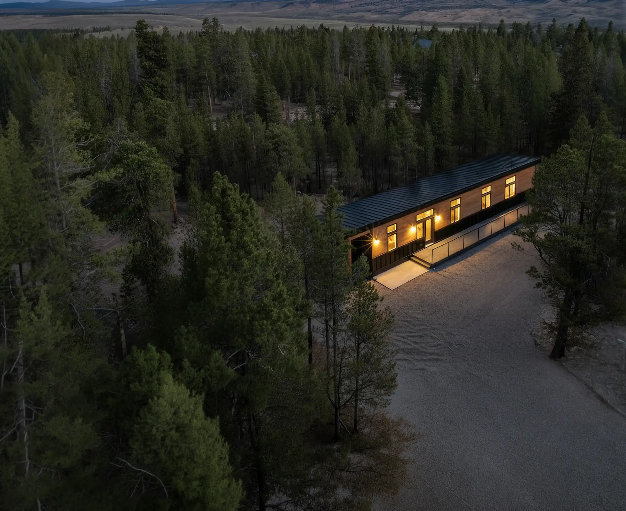 Aerial view of a long narrow home with dark metal roof nestled deep in a dense pine forest at dusk