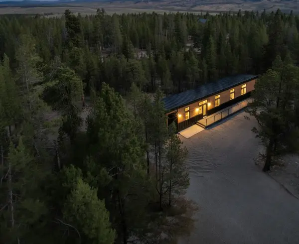 Aerial view of a long narrow home with dark metal roof nestled deep in a dense pine forest at dusk