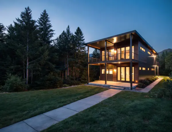 Two-story home with dark siding and large upper windows tucked into a lush green pine forest at night