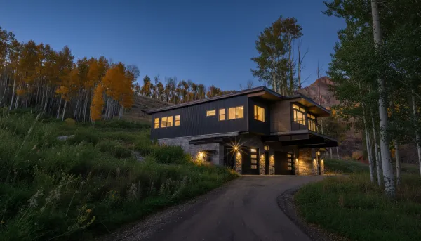Two-story dark modern home tucked into a hillside among golden aspens, with stone base and single garage