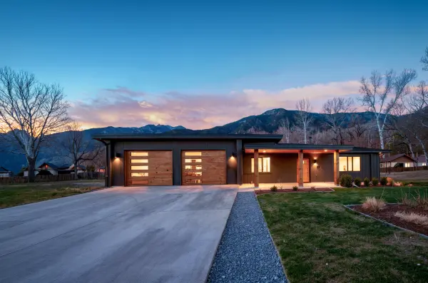Single-story ranch-style home with dark exterior, cedar-paneled garage doors, and wide driveway set against foothills at sunset