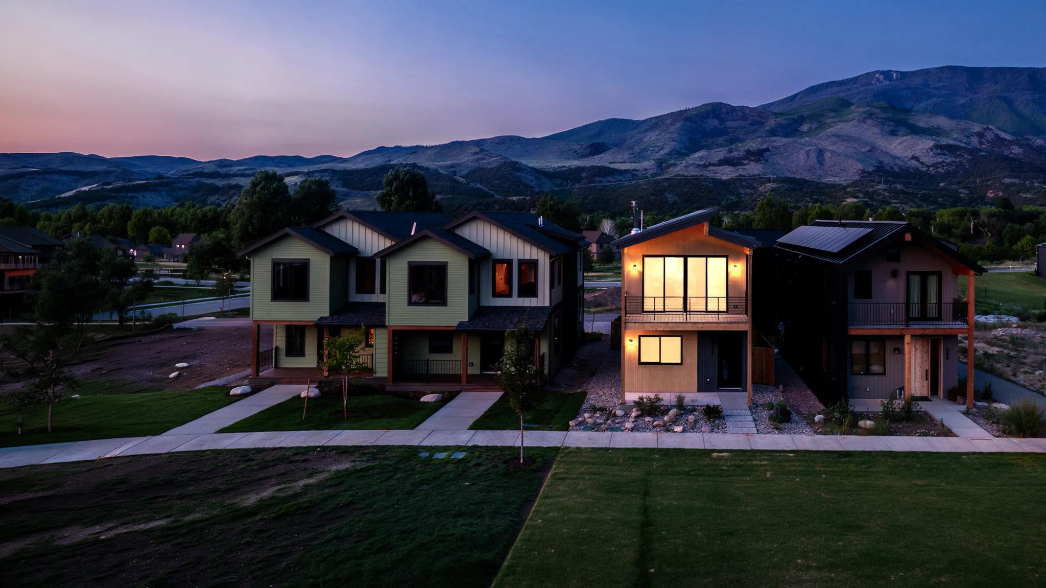 Three adjacent two-story modular homes with mixed wood and dark siding in a residential neighborhood at twilight, with rolling mountain foothills behind
