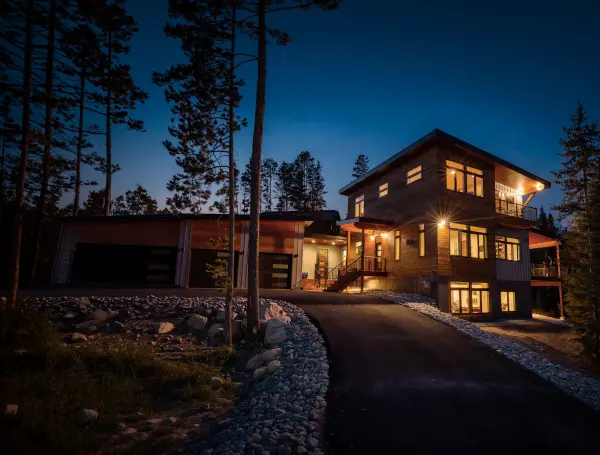 Large multi-level home with warm wood siding and two-car garage, set along a curved river-rock-lined driveway in a dense pine forest at night