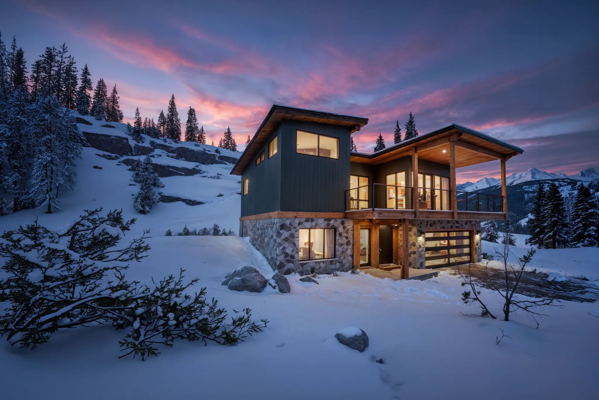 Two-story mountain home with dark metal upper level and stone masonry base, surrounded by deep snow with a pink alpine sunset behind
