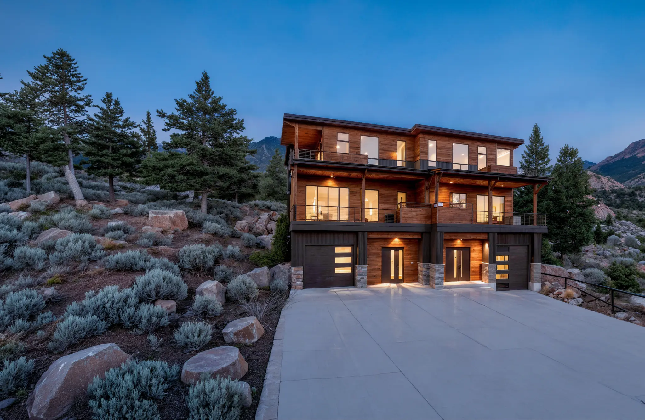 Three-story mountain home with cedar siding and large windows on a boulder-lined hillside, photographed at twilight with rocky peaks behind
