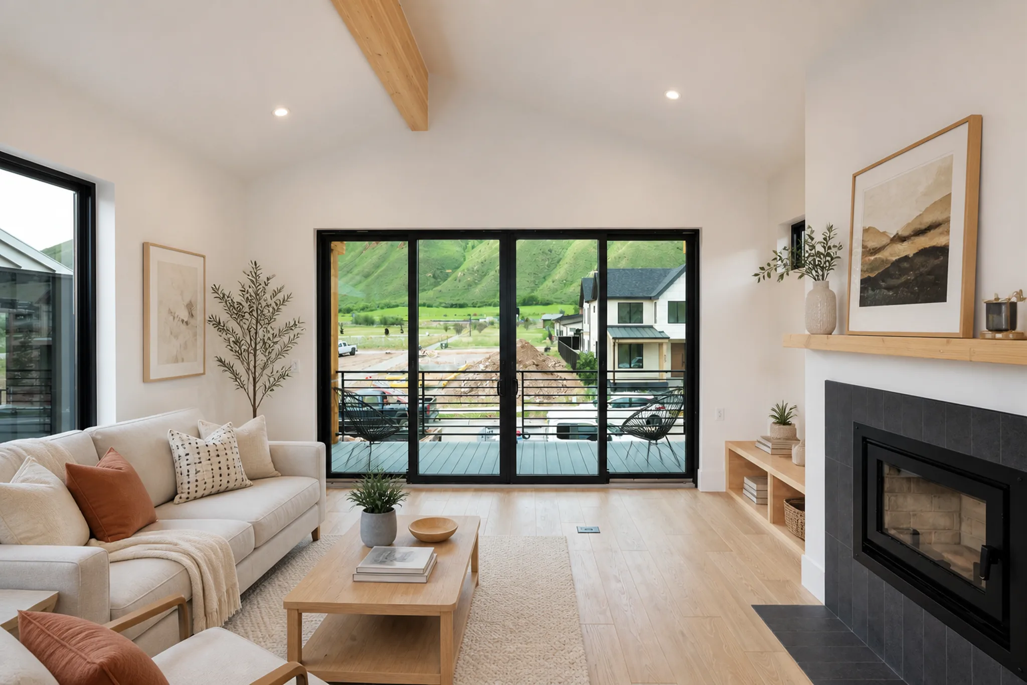 Bright living room with vaulted ceiling, exposed wood beam, cream sofa, black-framed sliding glass doors opening to balcony with green mountain views, and gas fireplace