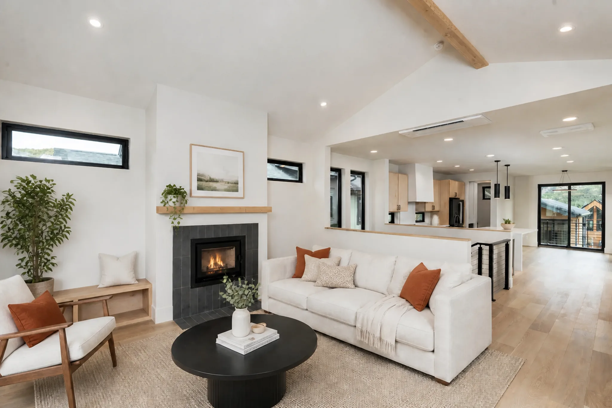 Open-plan living area with vaulted ceiling, exposed beam, cream sofa, gas fireplace, and open view into kitchen with light wood cabinetry, island, and pendant lights