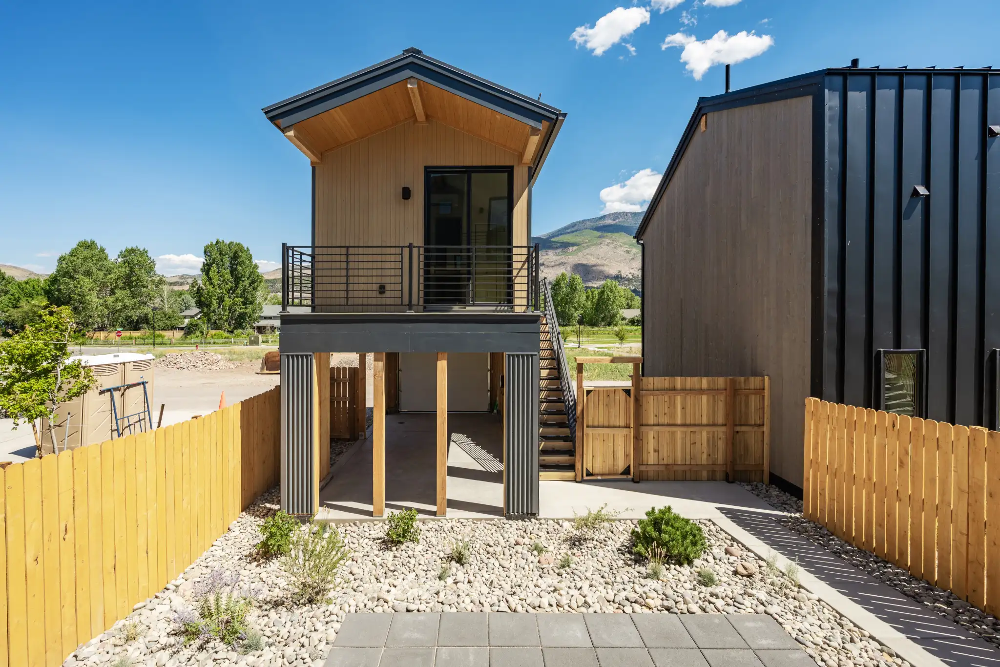 Rear exterior of two-story home with wood and corrugated metal siding, upper balcony with metal railing, exterior staircase, gravel xeriscaping, and mountain backdrop