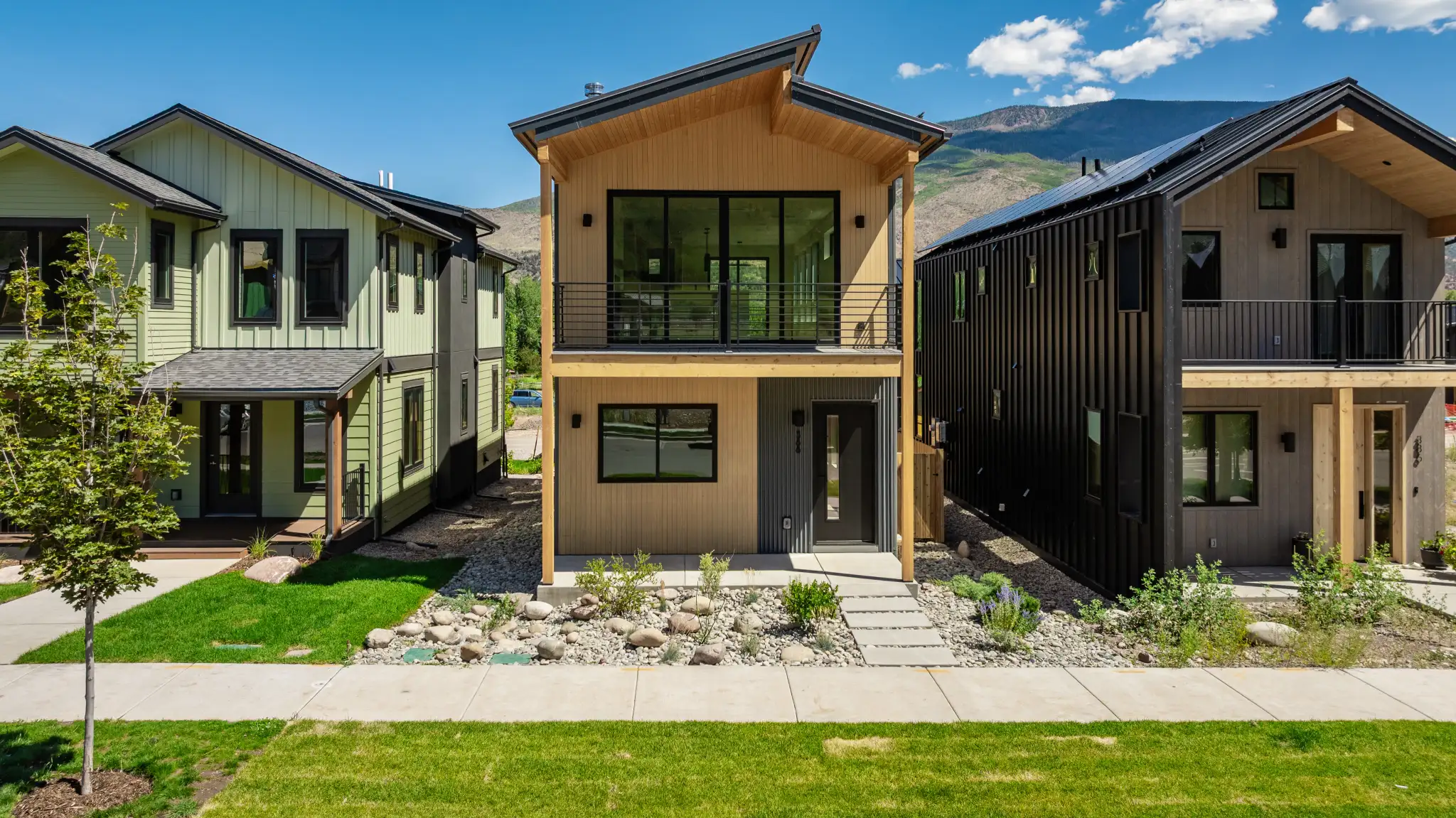 Front exterior of two-story home with tan stucco lower level, warm wood upper story, black trim, upper balcony, and mountain backdrop