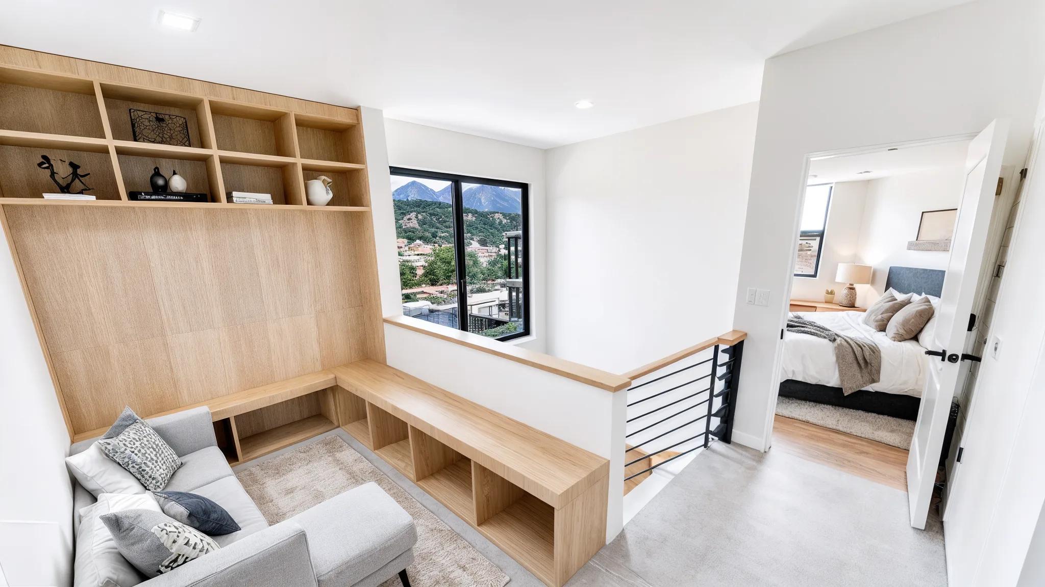 Upper loft landing with built-in wood shelving and desk, gray sofa, black-framed window with mountain view, and open doorway into bedroom