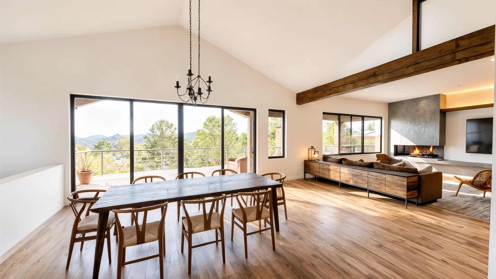 Open dining and living area with vaulted ceiling, exposed wood beams, large dark wood dining table with wishbone chairs, black chandelier, and floor-to-ceiling windows with mountain and tree views
