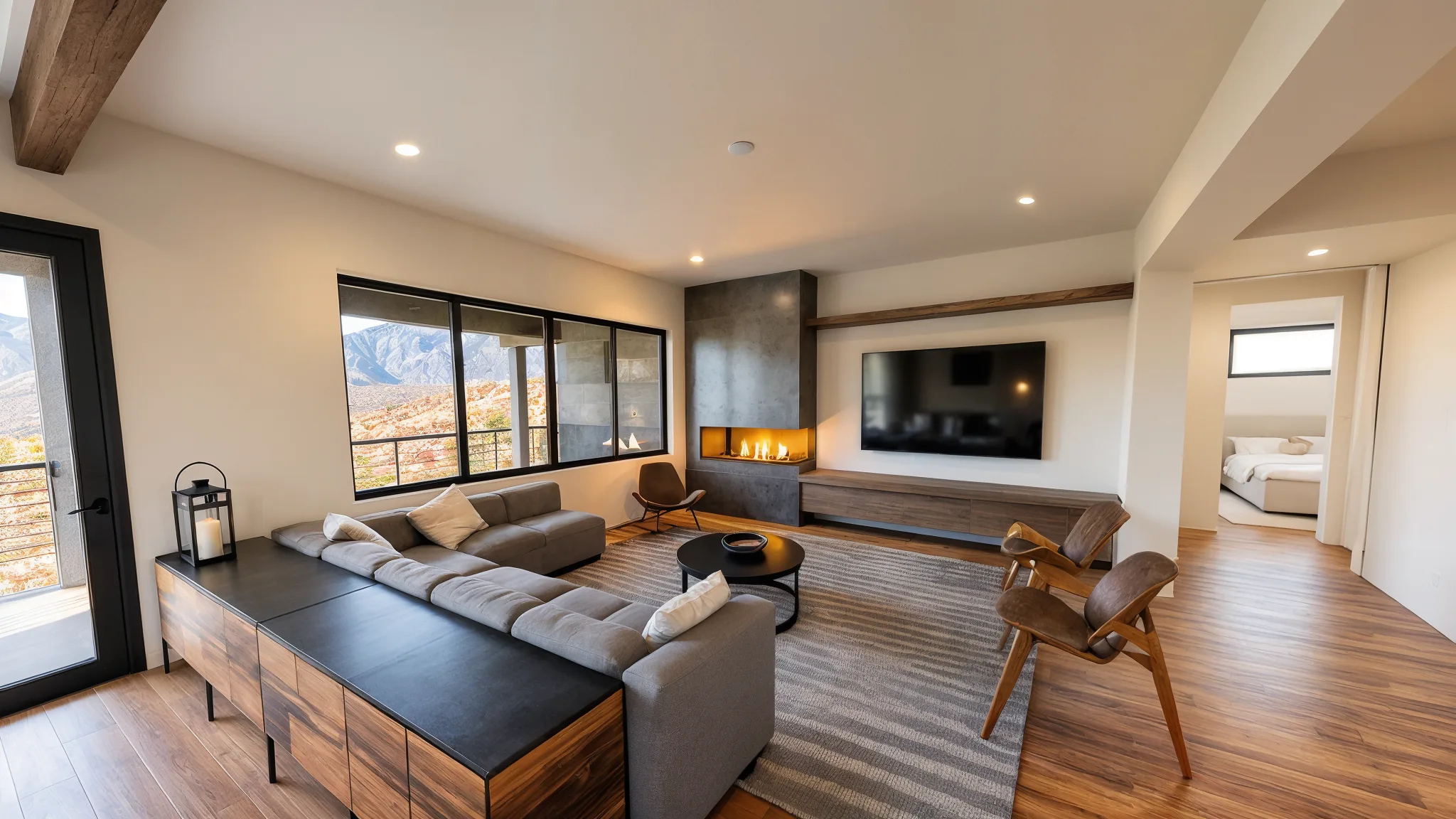 Modern living room with gray sectional sofa, striped rug, corner gas fireplace with dark metal surround, wall-mounted TV, exposed wood beam, and large windows with mountain views