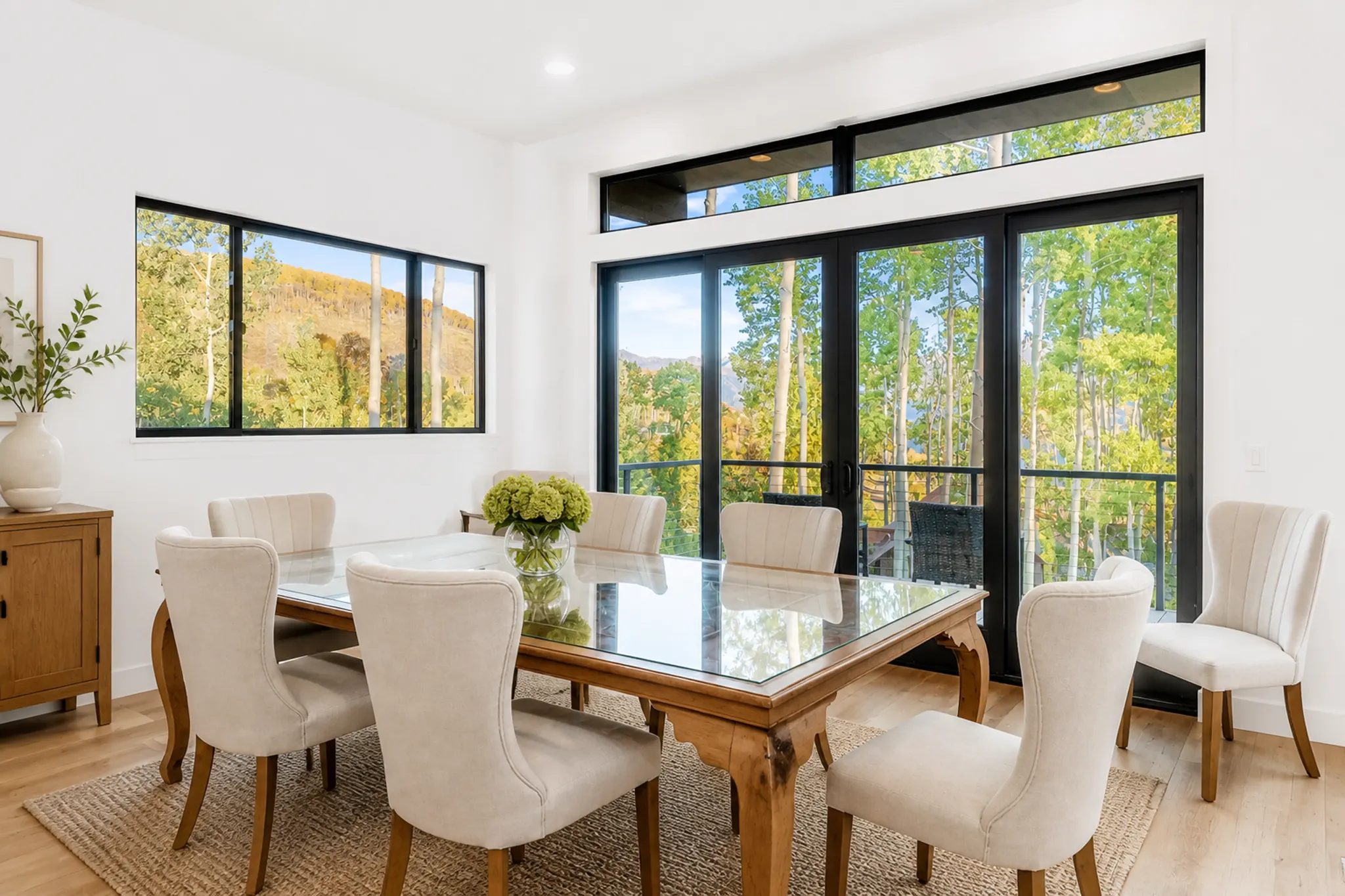 Dining room with wood-framed glass dining table, cream wingback chairs, black-framed floor-to-ceiling windows with fall aspen tree views, and light wood floors