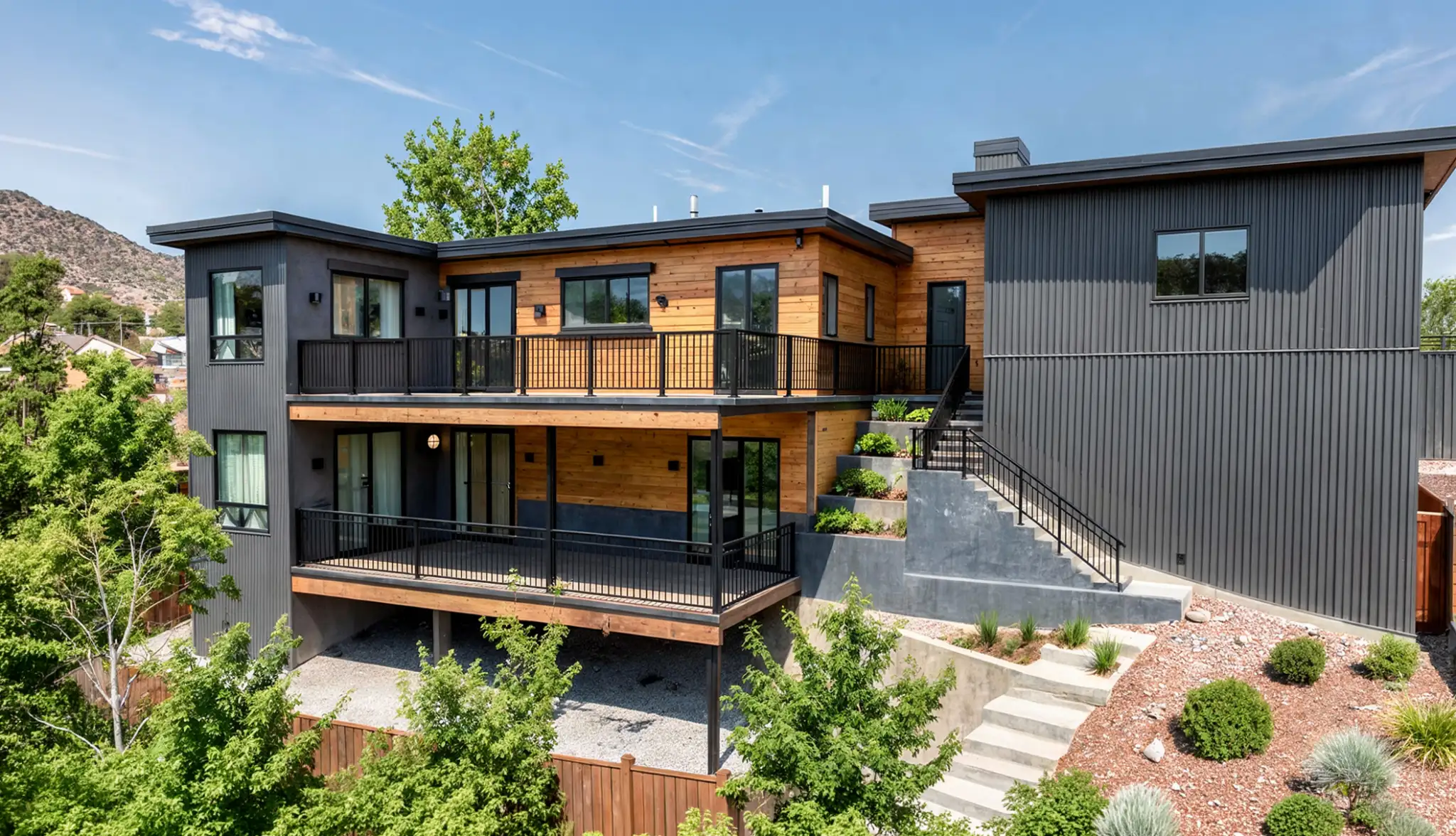 Two-story home with dark gray vertical siding, warm cedar upper-level cladding, two-level balconies with black metal railings, exterior staircase, and terraced native plant landscaping