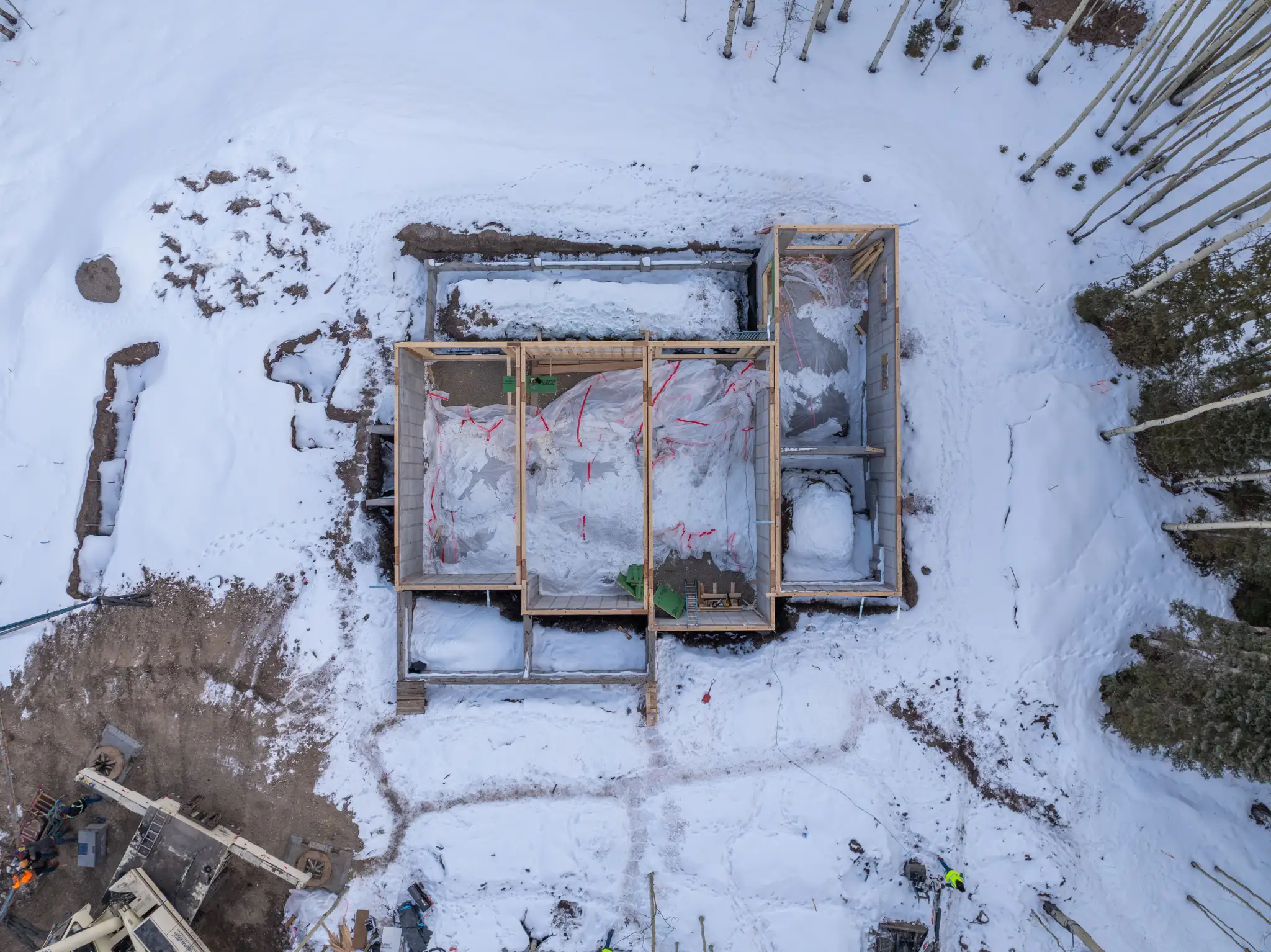 Overhead aerial of snow-covered foundation framing with bare aspen trees and construction workers