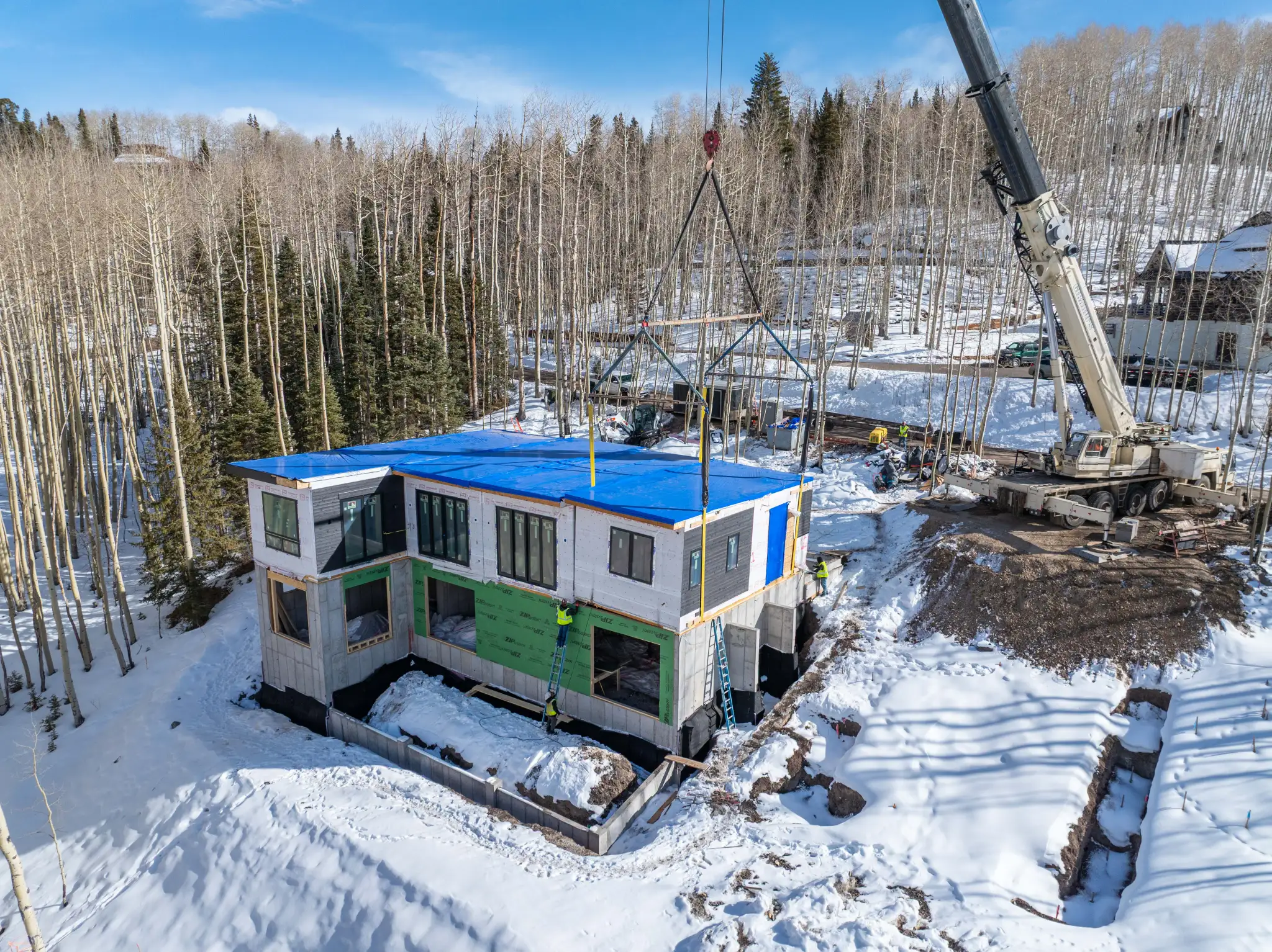 Aerial view of modular home module being craned onto snowy foundation amid bare aspen trees, with construction crew visible and blue temporary roof tarp