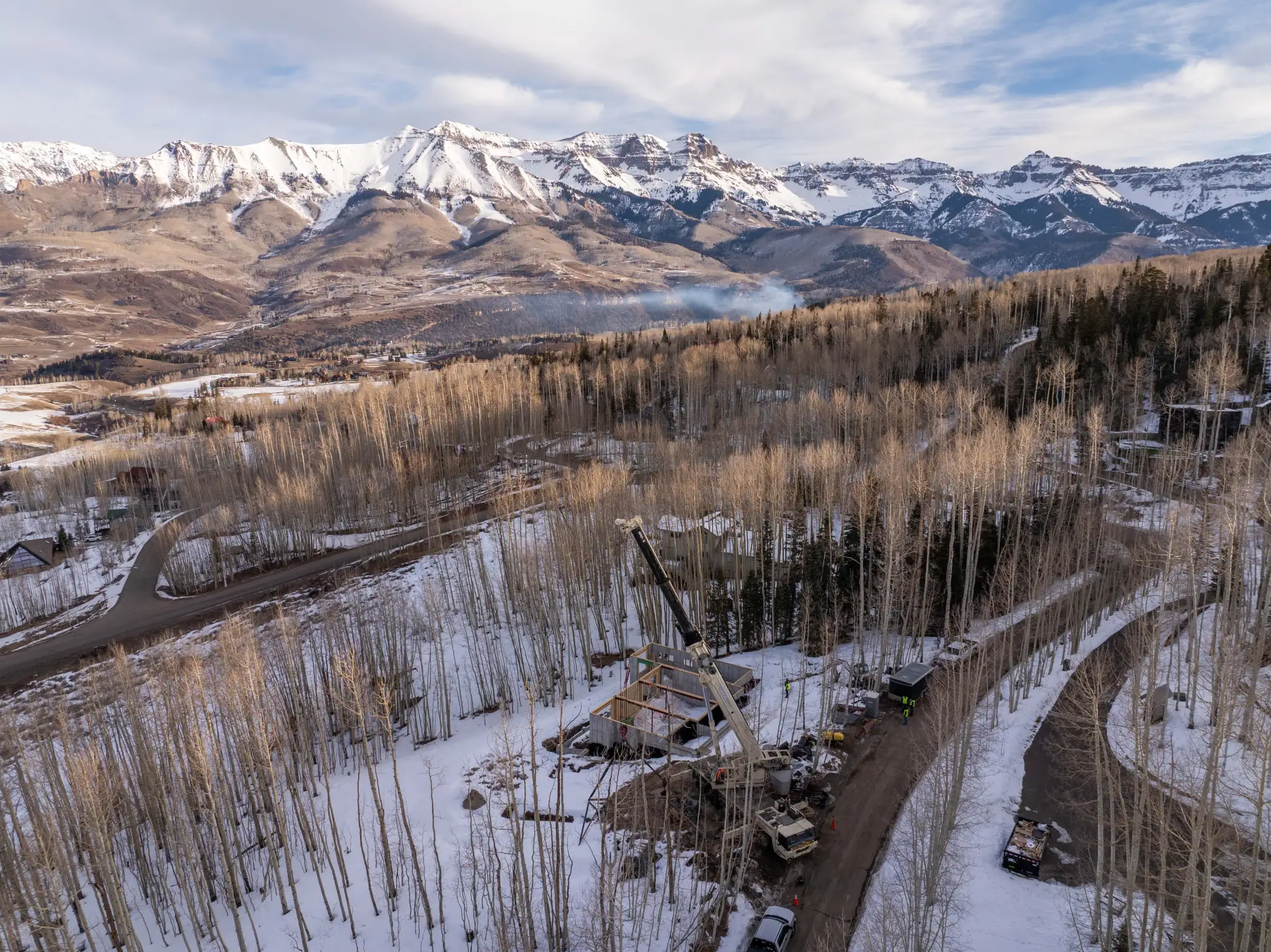 Wide aerial view of construction site in snowy aspen forest with crane, sweeping mountain valley, and snow-capped peaks in background