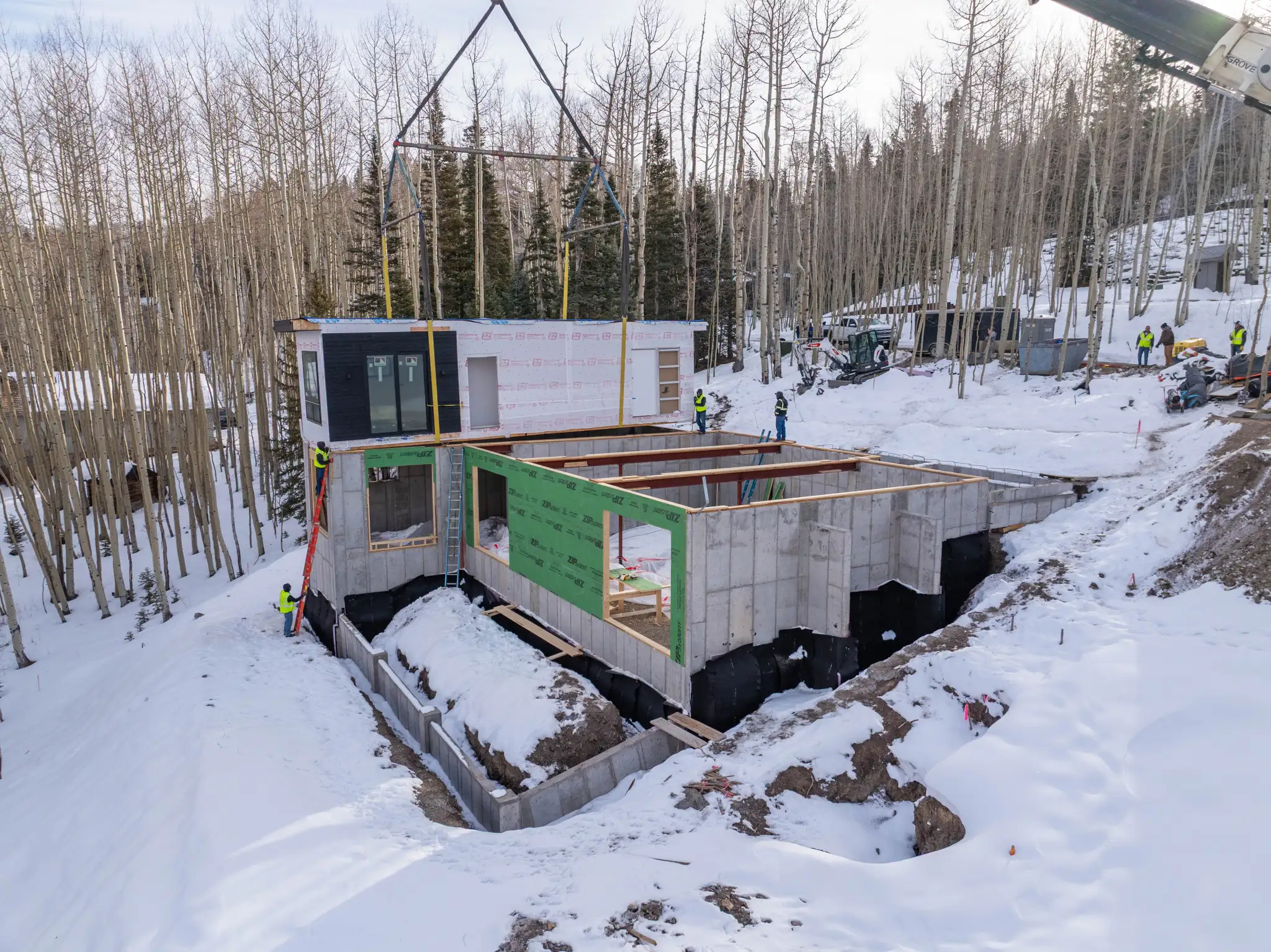 Side view of modular home module being craned onto hillside foundation with crew in hi-vis vests and bare aspens in winter landscape
