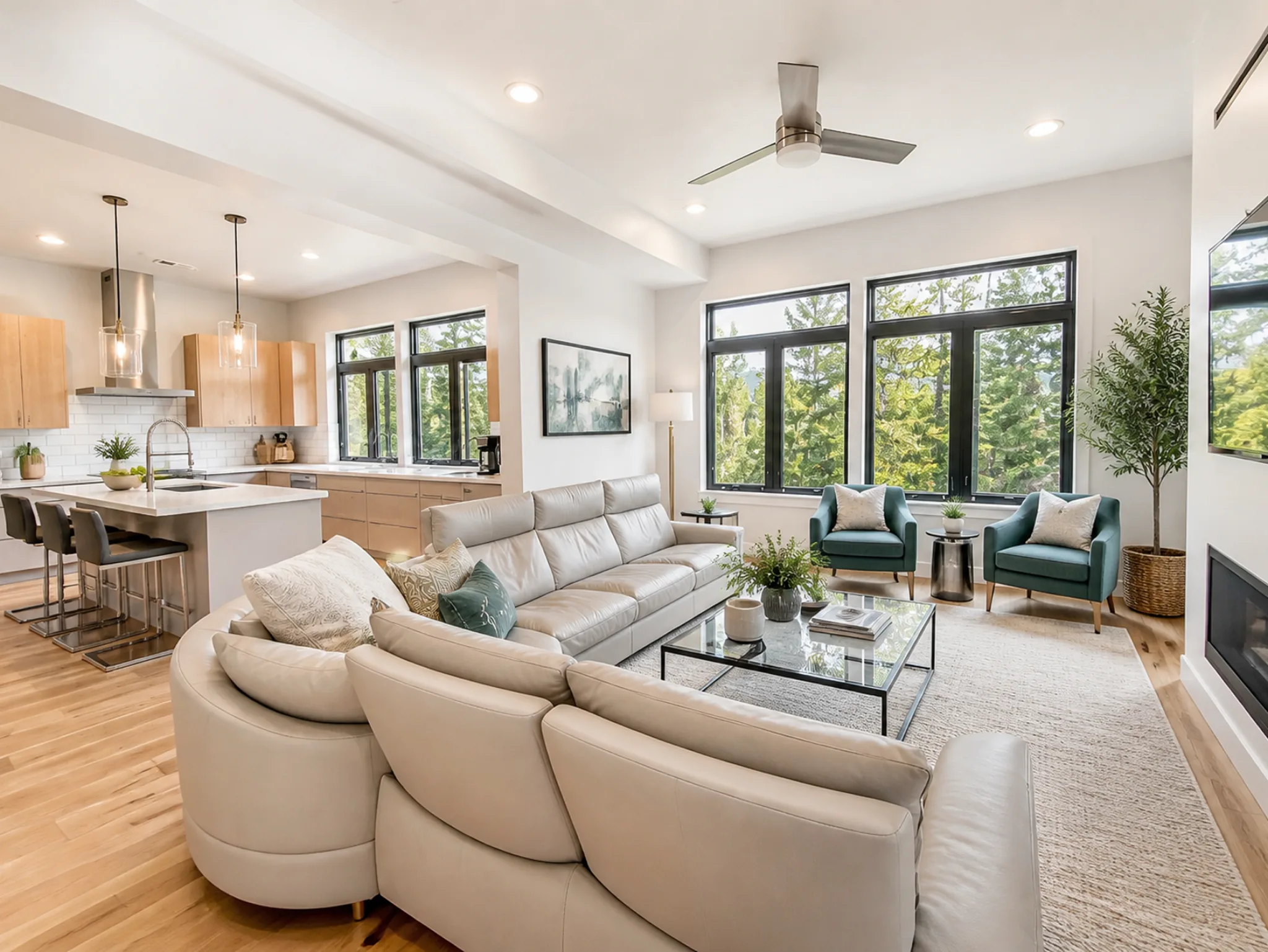 Open-plan living area with large cream curved sectional, two teal accent chairs, glass coffee table, gas fireplace, and floor-to-ceiling black-framed windows with pine tree views