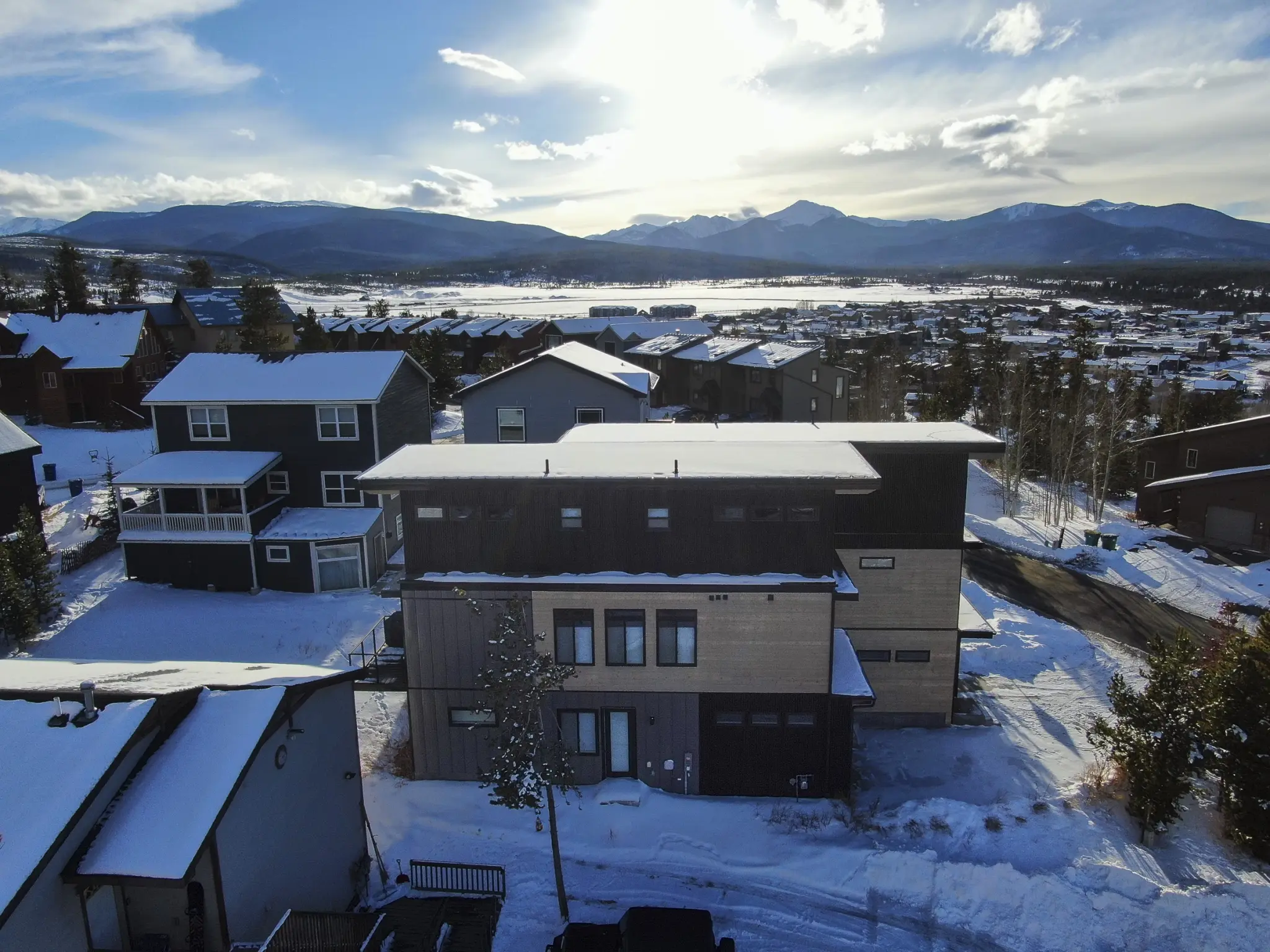 Aerial view of three-story dark home with cedar accent section, snow-covered flat roof, snowy residential neighborhood, mountain range, and frozen lake in background