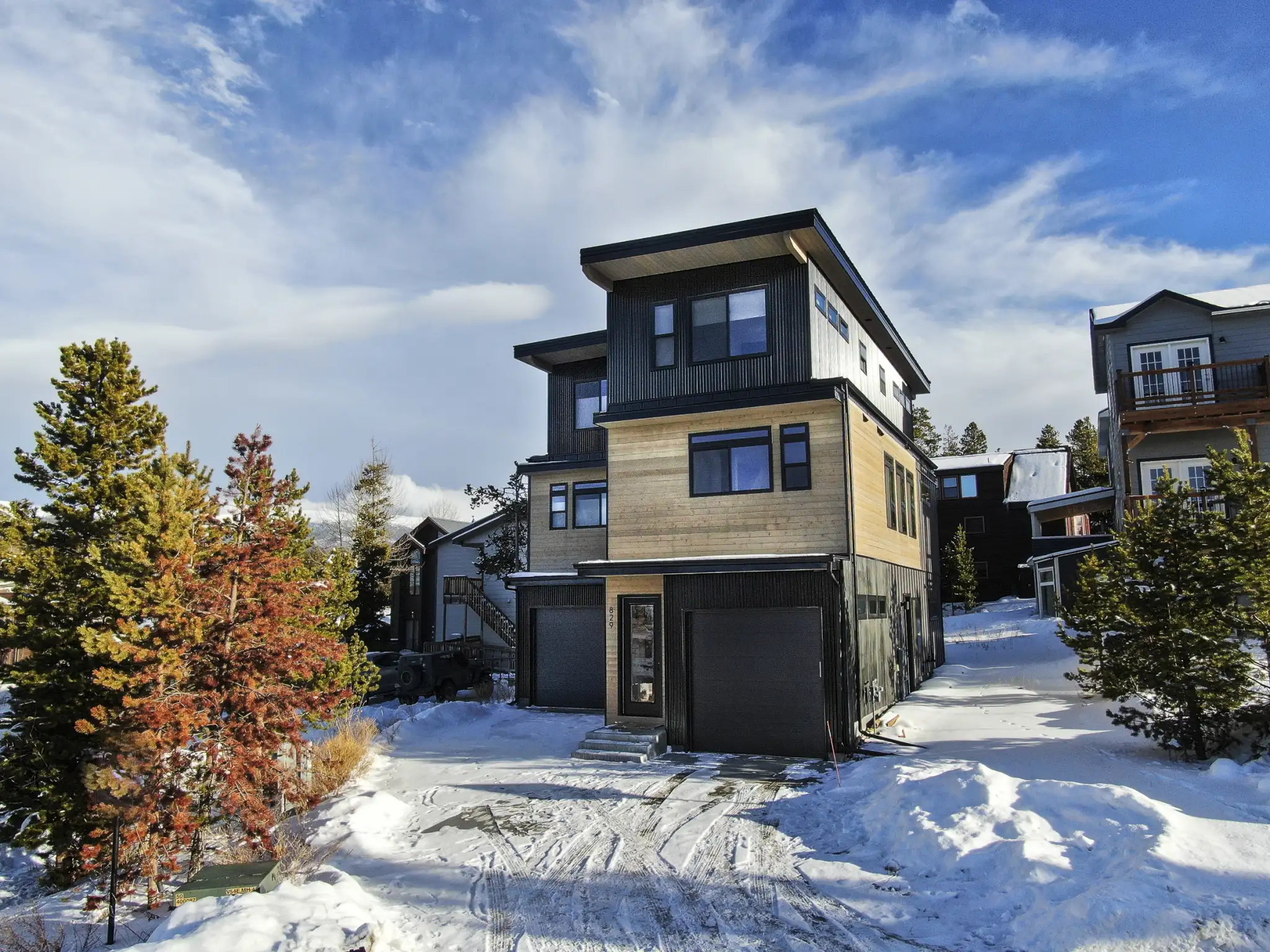 Three-story home with dark vertical corrugated metal siding, warm cedar middle-level cladding, double-car garage, snowy setting with pine trees and neighboring homes