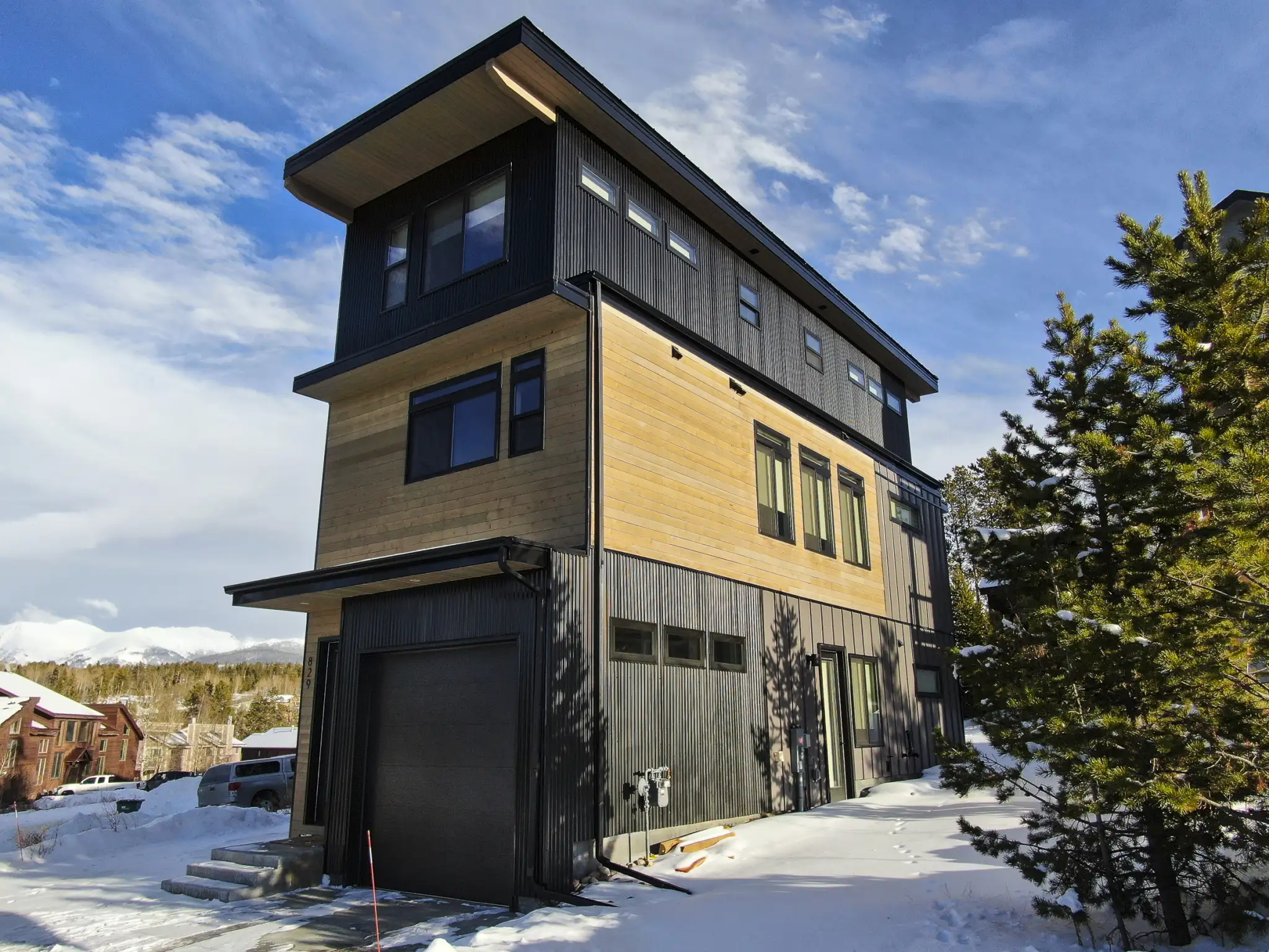 Three-story home near completion with dark upper and lower metal siding, warm cedar middle section, single-car garage, and snowy setting with mountain backdrop