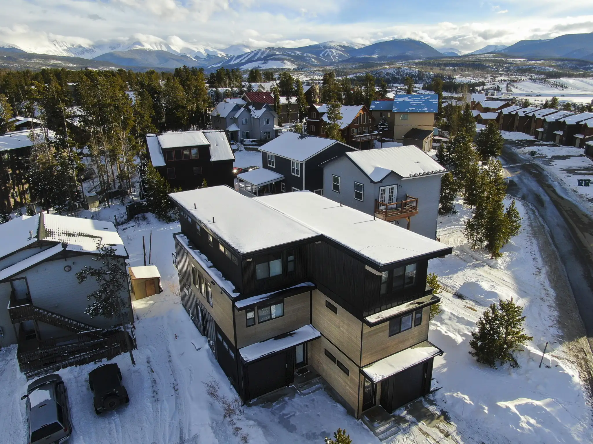 Aerial view of completed three-story home with dark metal siding and cedar middle section, snow-covered flat roof, neighboring homes, pine trees, and distant snow-capped mountains